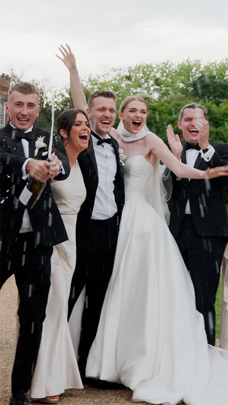A group of five people celebrating outdoors, dressed in formal wedding attire, with happy expressions and raised arms.