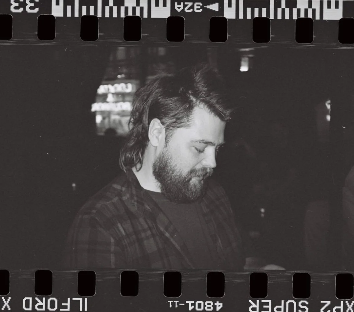 A black and white photo of a man with long hair and a beard, looking down with a calm expression in a dimly lit setting.