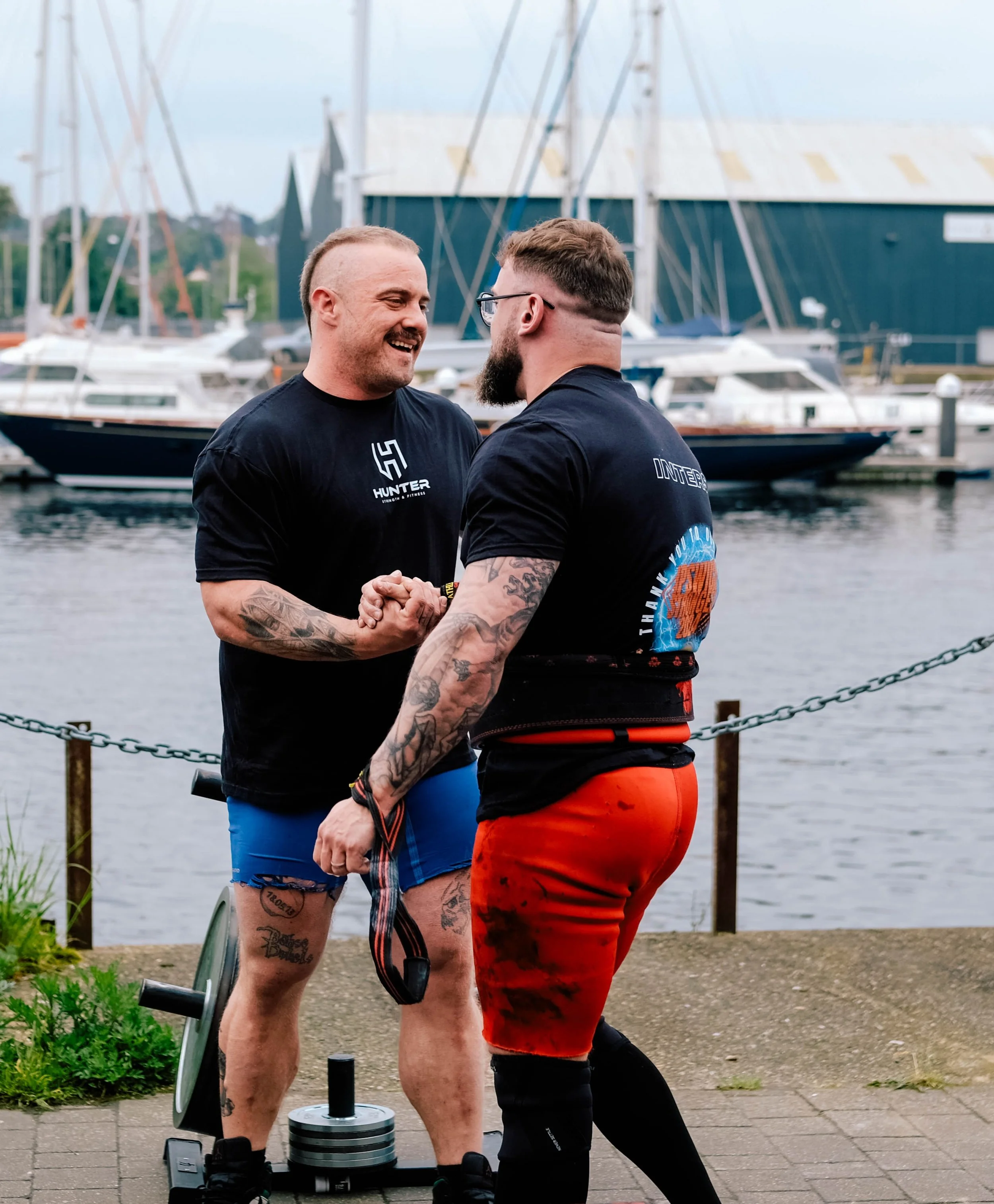 Two men are smiling and shaking hands near a waterfront with boats, including yachts and sailboats, in the background.