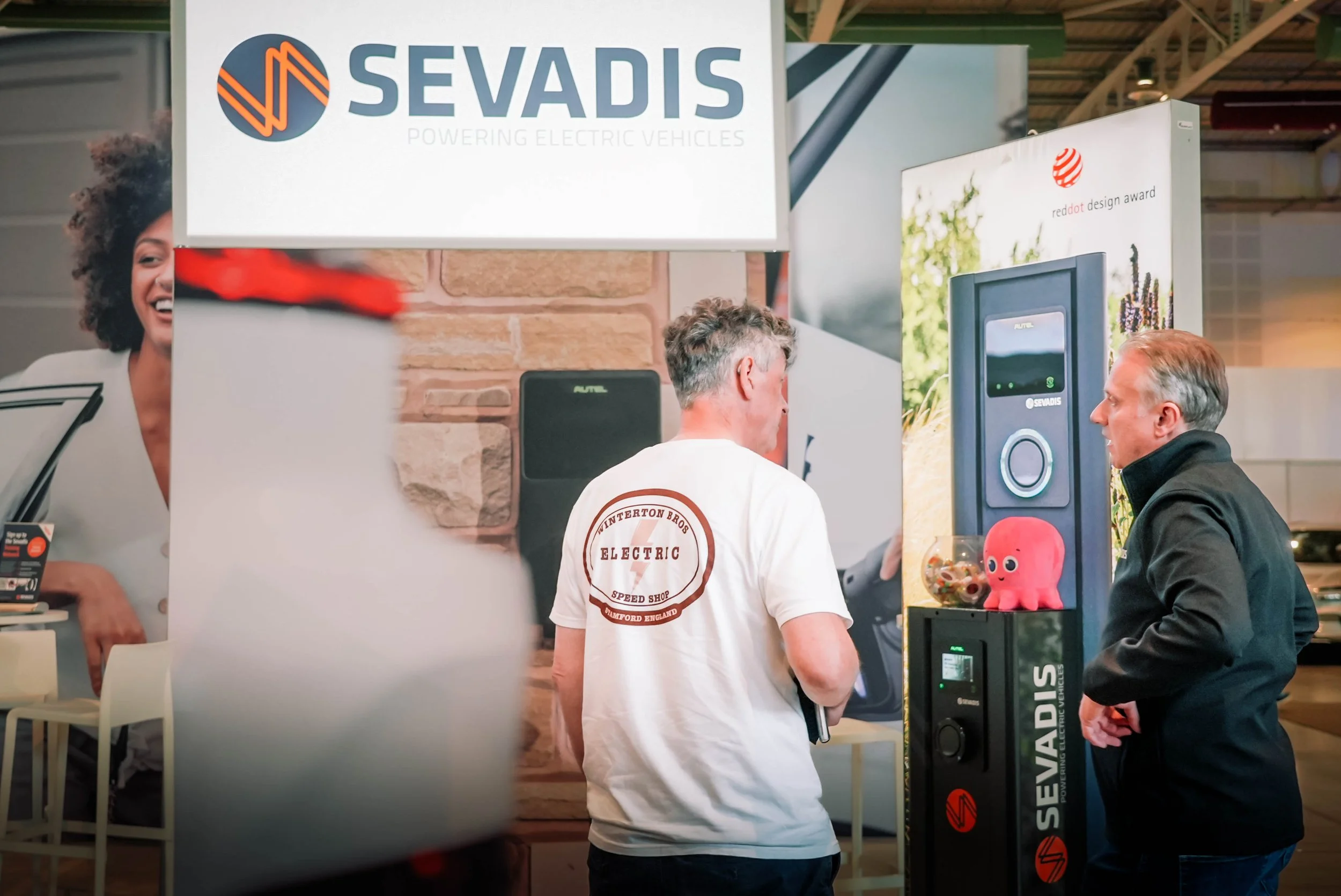 People at an exhibition booth with a sign reading 'Sevadis Powering Electric Vehicles'. Two men are engaged in conversation; one wears a white T-shirt with a logo reading 'Winterton Bros Electric Speed Shop' from Radford, England, and the other wears