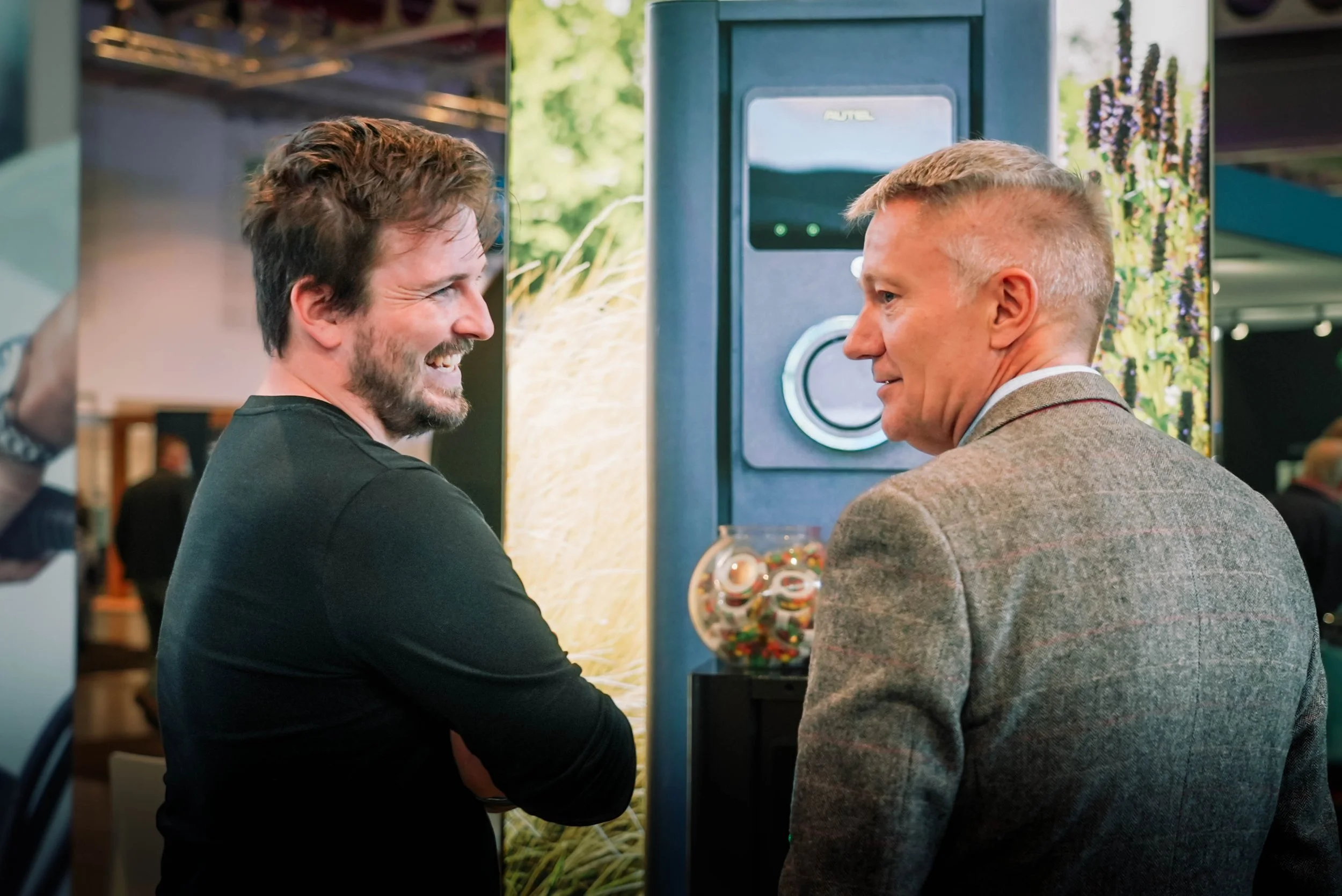 Two men, one with brown hair and beard and another with short blonde hair, are smiling and standing close together. They are facing each other and appear to be enjoying a conversation. They are standing in front of a vending machine filled with color