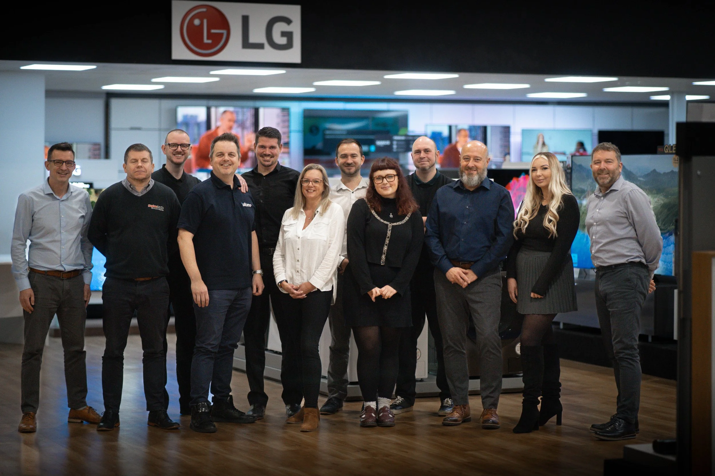 Group of 12 professional adults, men and women, standing inside an electronics store with LG TVs and monitors in the background.