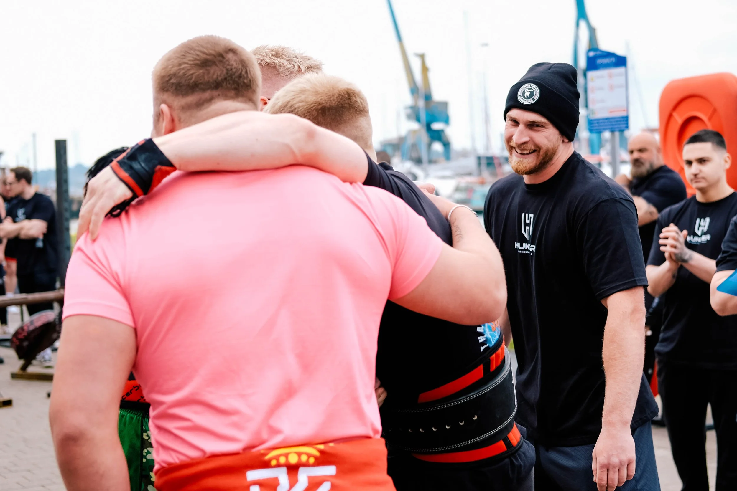 A group of men are gathered outdoors near a marina, engaging in a group hug and smiling, some are wearing athletic clothing and beanies.
