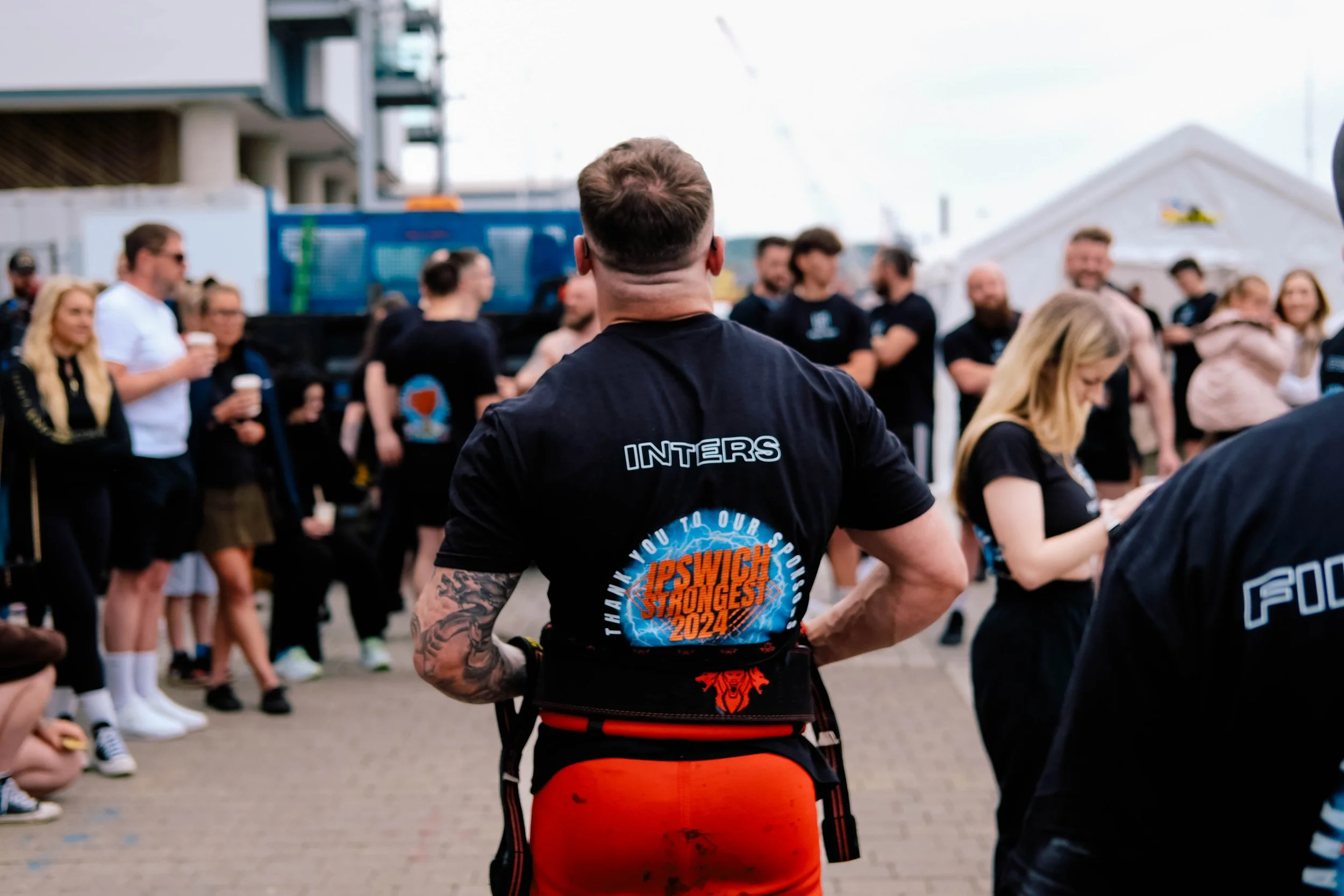 Man with tattoos on arms wearing black shirt with text and logo on the back, standing in front of a group of people outdoors, participating in or observing a Strongman competition or event.