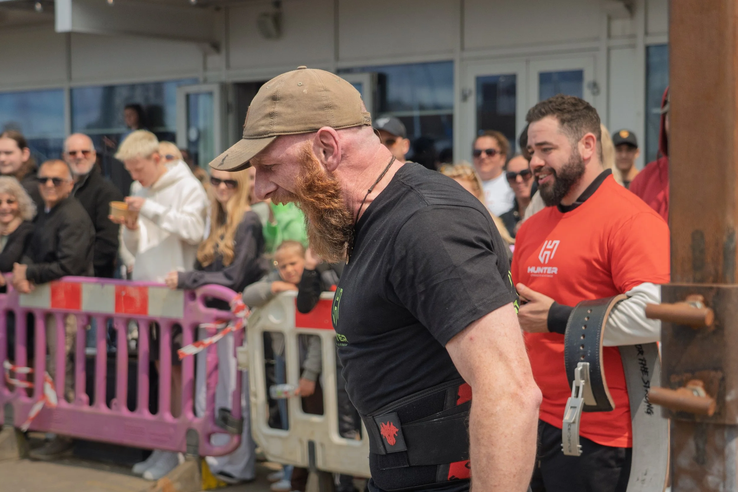 A group of people gathered outdoors, with two men in the foreground preparing for a strength or fitness event, surrounded by spectators.