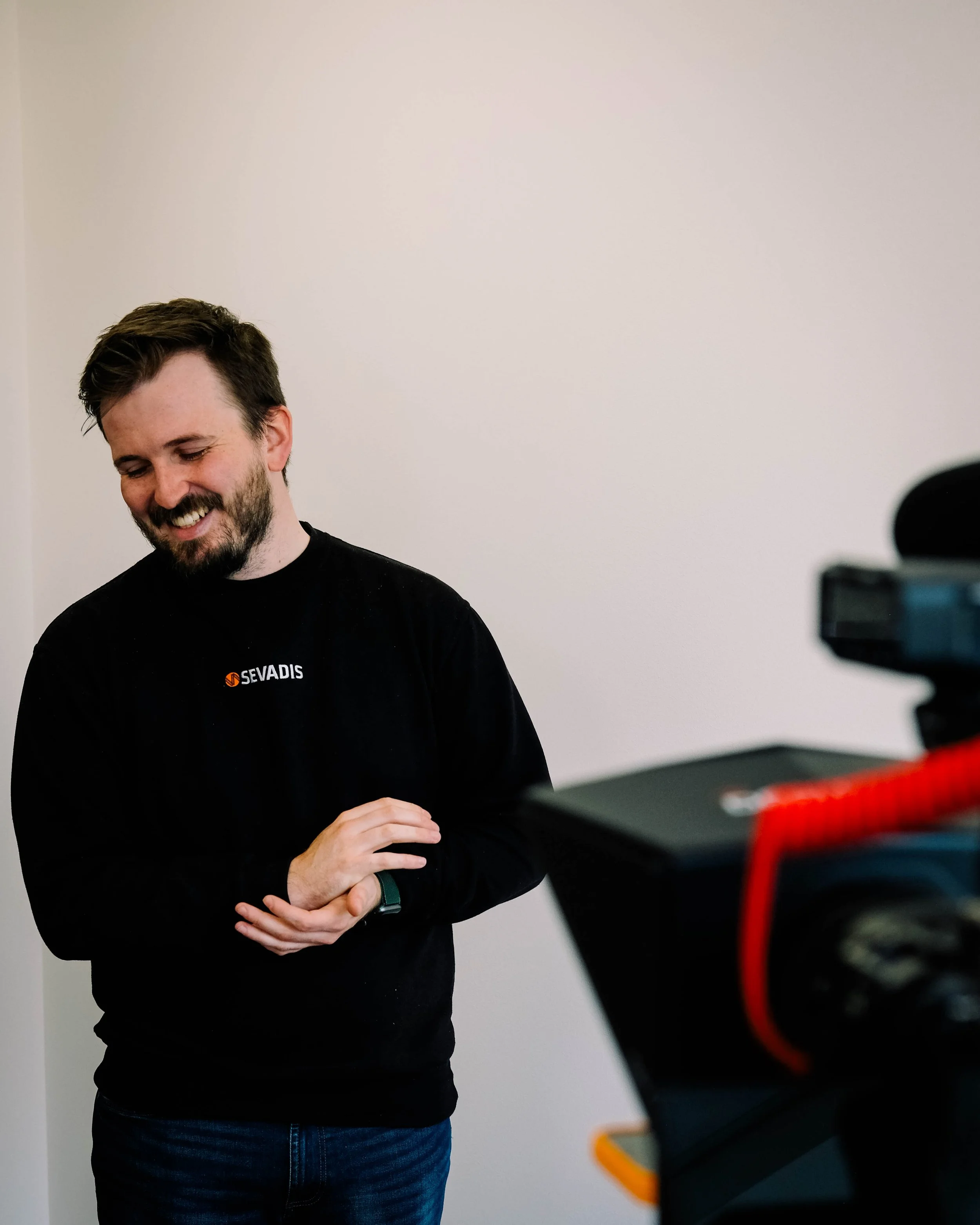 A man with a beard and short dark hair is smiling and looking down, standing in a room with plain white walls. He is wearing a black long-sleeve shirt with a logo reading 'SEVADIS' on the chest and a smartwatch on his left wrist. Part of a camera or 