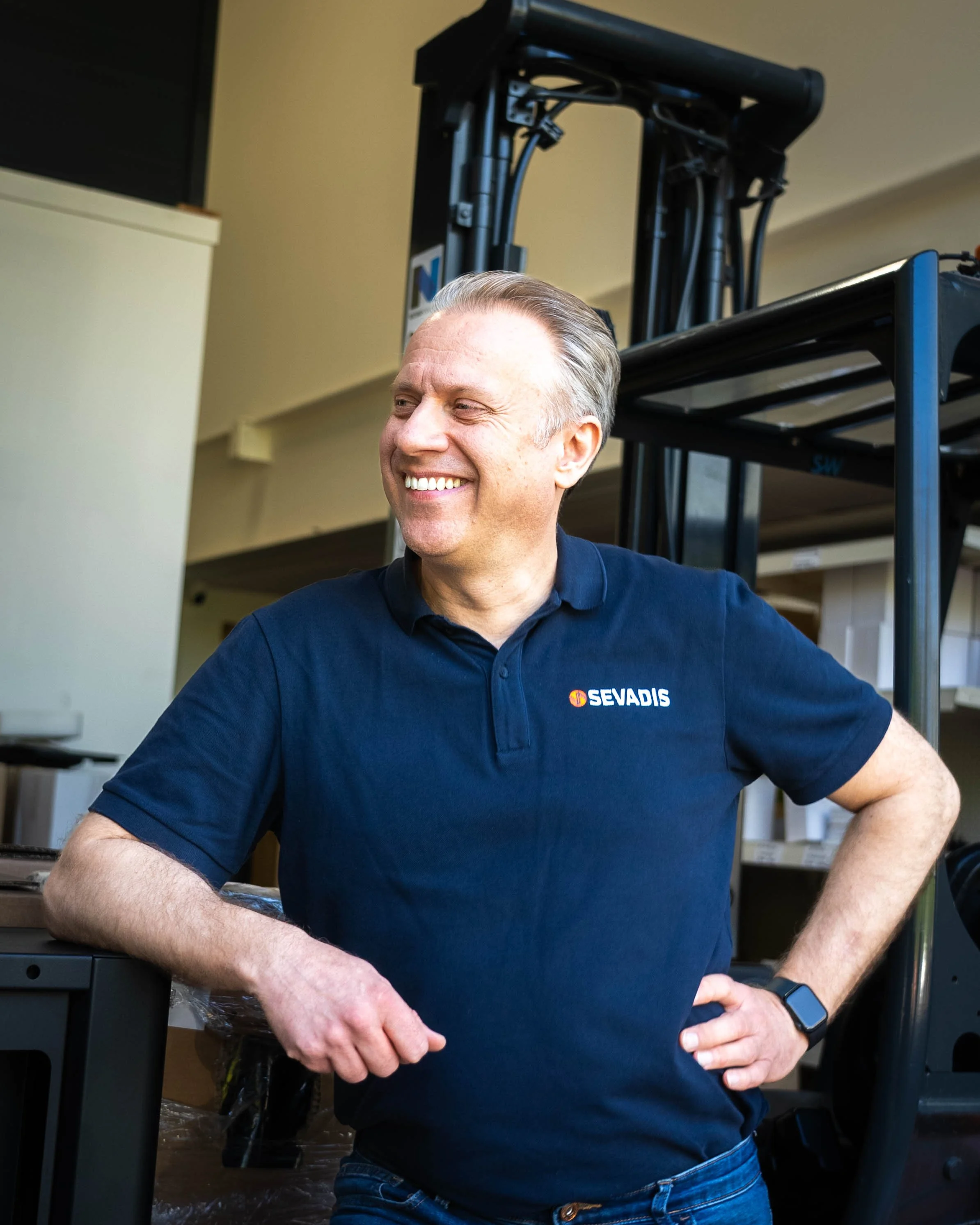 Smiling man wearing a navy blue polo shirt with a Sevadis logo, standing indoors with shelves and equipment behind him.
