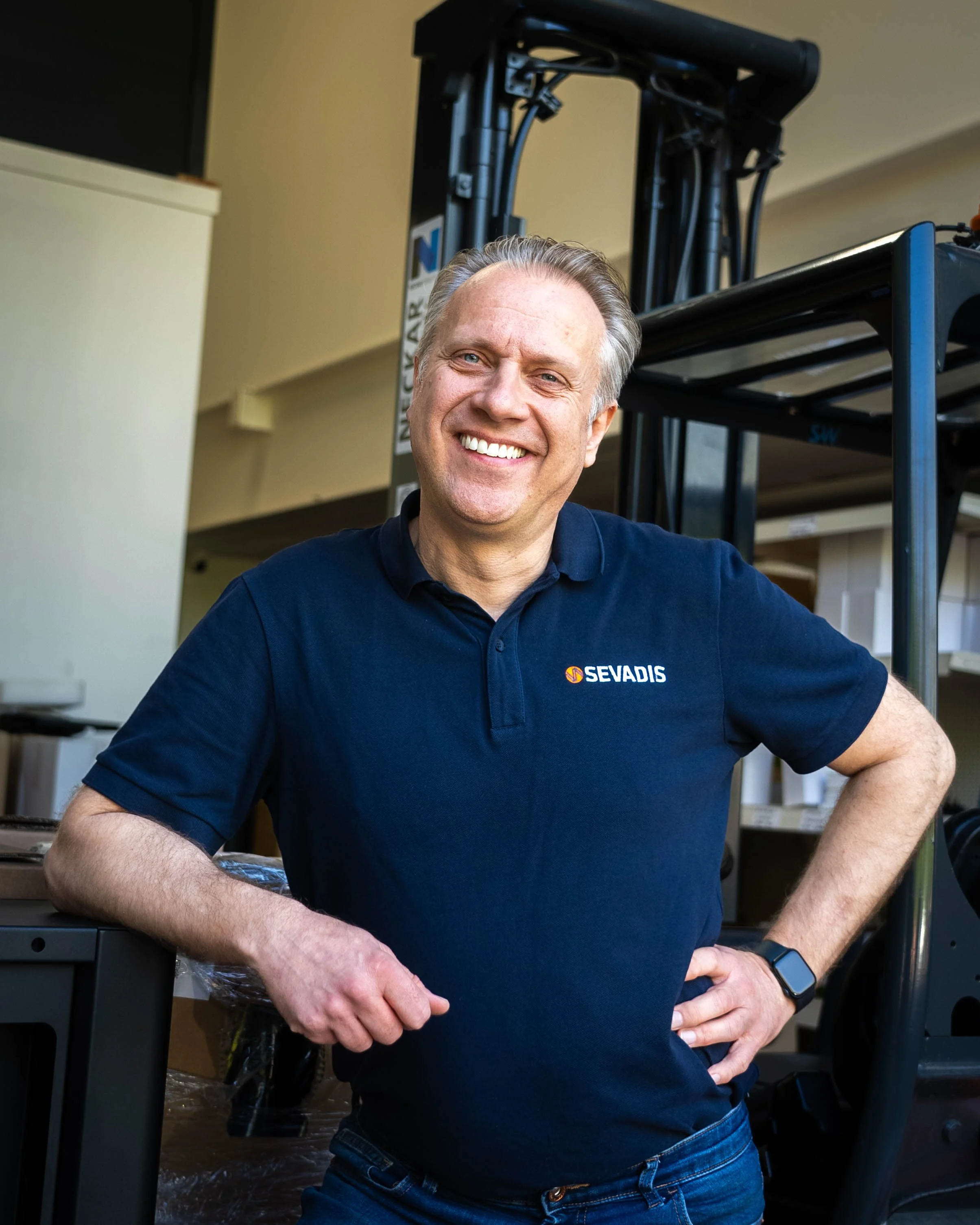 A smiling man in a navy blue polo shirt stands with one hand on his hip and the other resting on a black work table. He has short, gray hair and wears a black smartwatch. The polo shirt has a logo that says 'SEVADIS' on it. The background shows vario