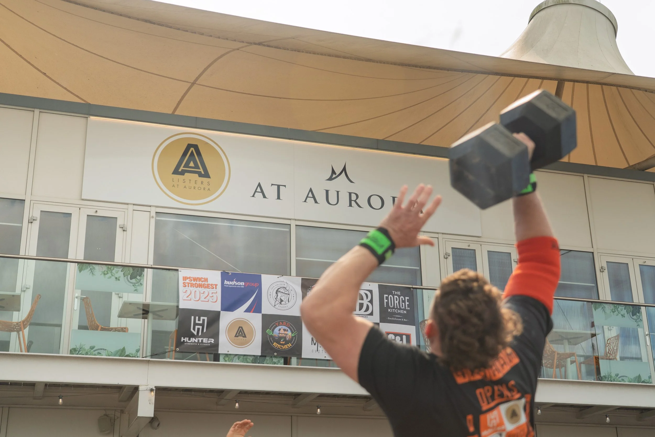 Person holding a hand in the air and raising an object, with a building and banners behind them, at an outdoor event near the Aurora Pavilion.