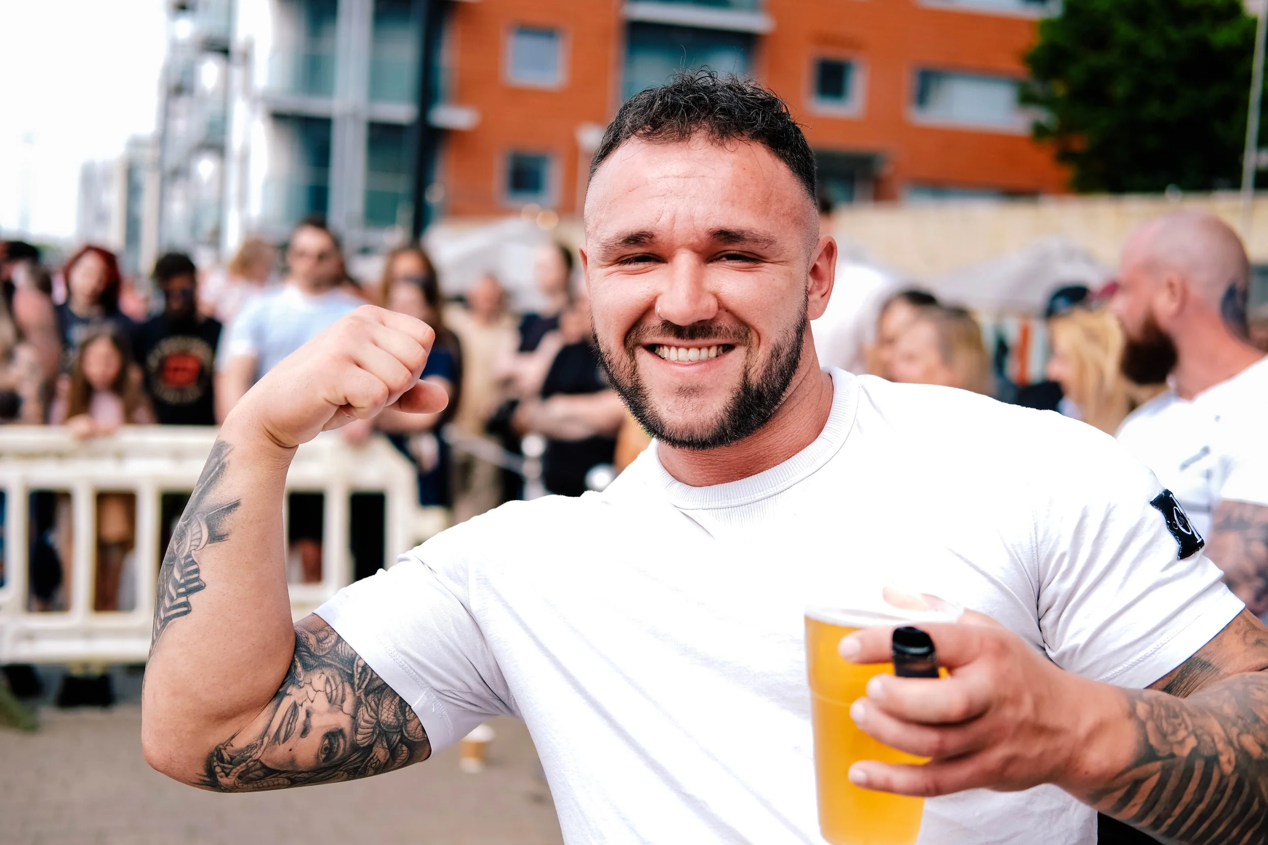 A smiling man wearing a white t-shirt and holding a yellow drink, flexing his bicep outdoors at a crowded event.