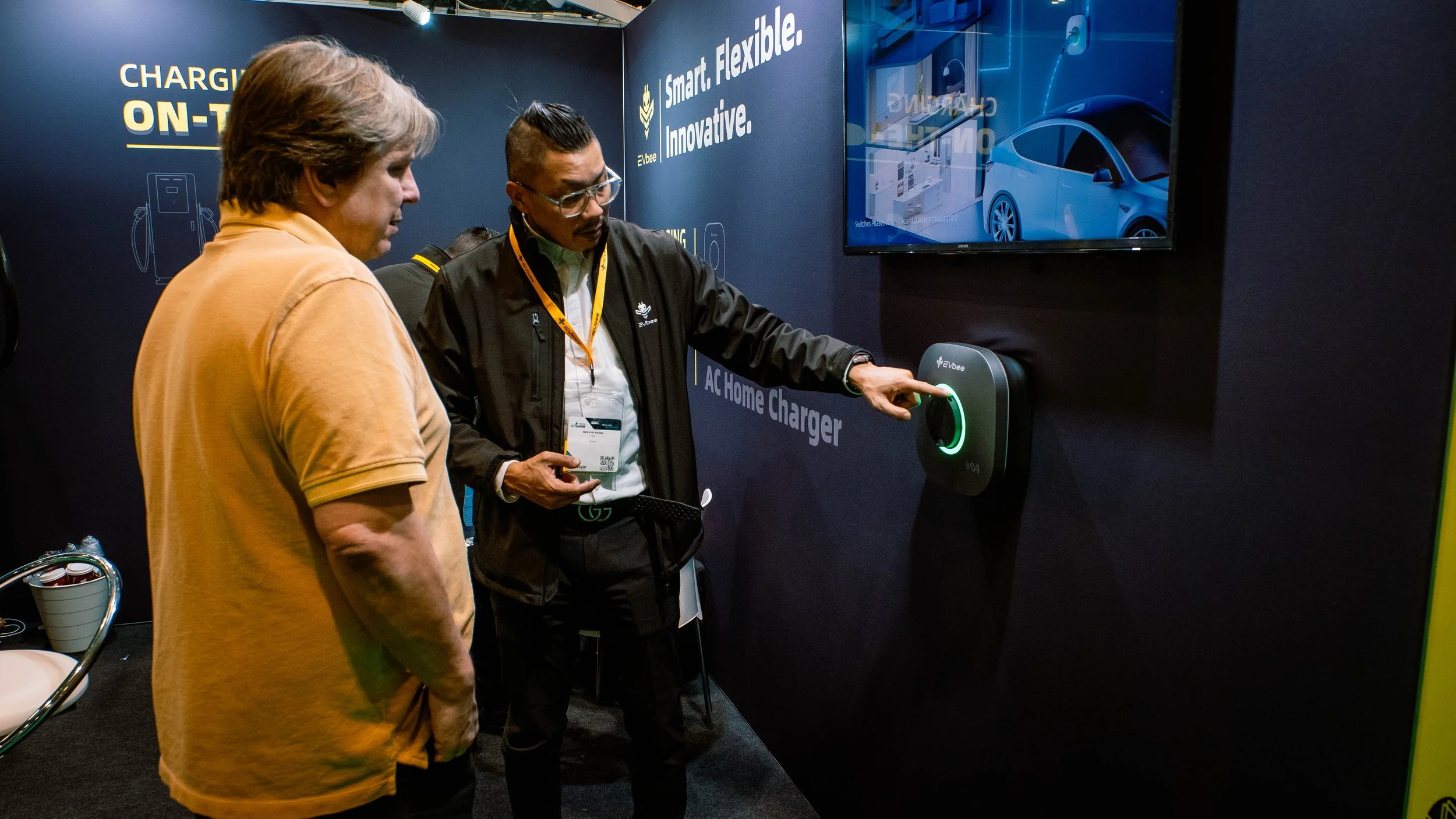 Two men are at an exhibition demonstrating an electric vehicle charger. One is pressing the charger while the other looks on. A large screen behind displays a blue car, and a promotional poster promoting smart, flexible, innovative charging solutions
