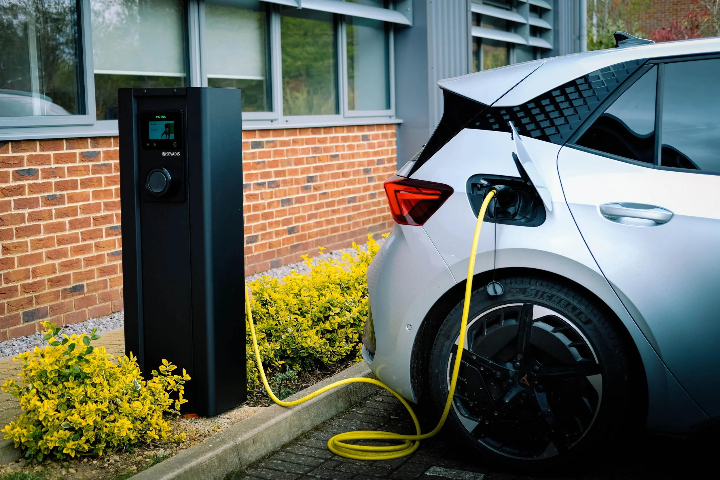 A white electric car is plugged into a charging station outside a building with large windows and brick wall. Bright yellow charging cable connected to the car, surrounded by green plants and yellow flowers.