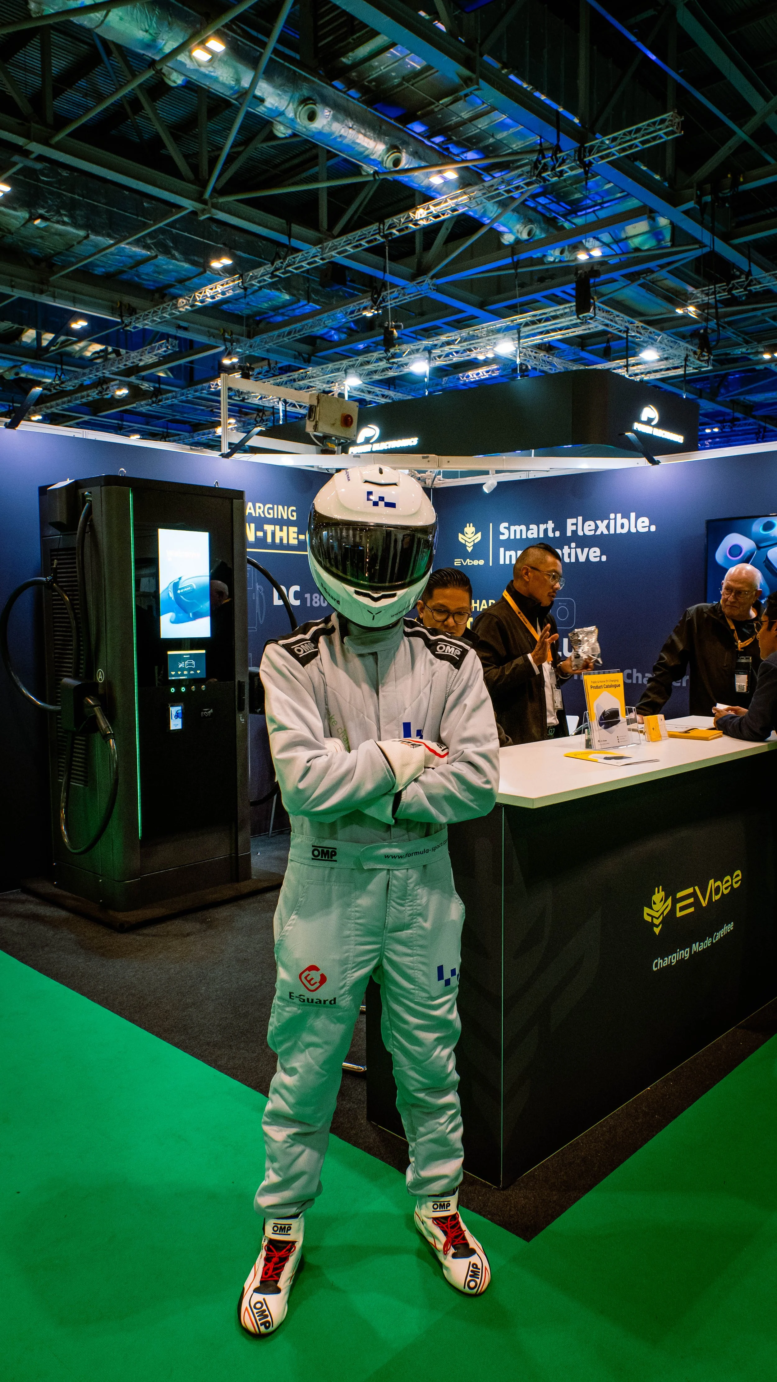 Person in racing suit with helmet standing in front of a booth at an exhibition.