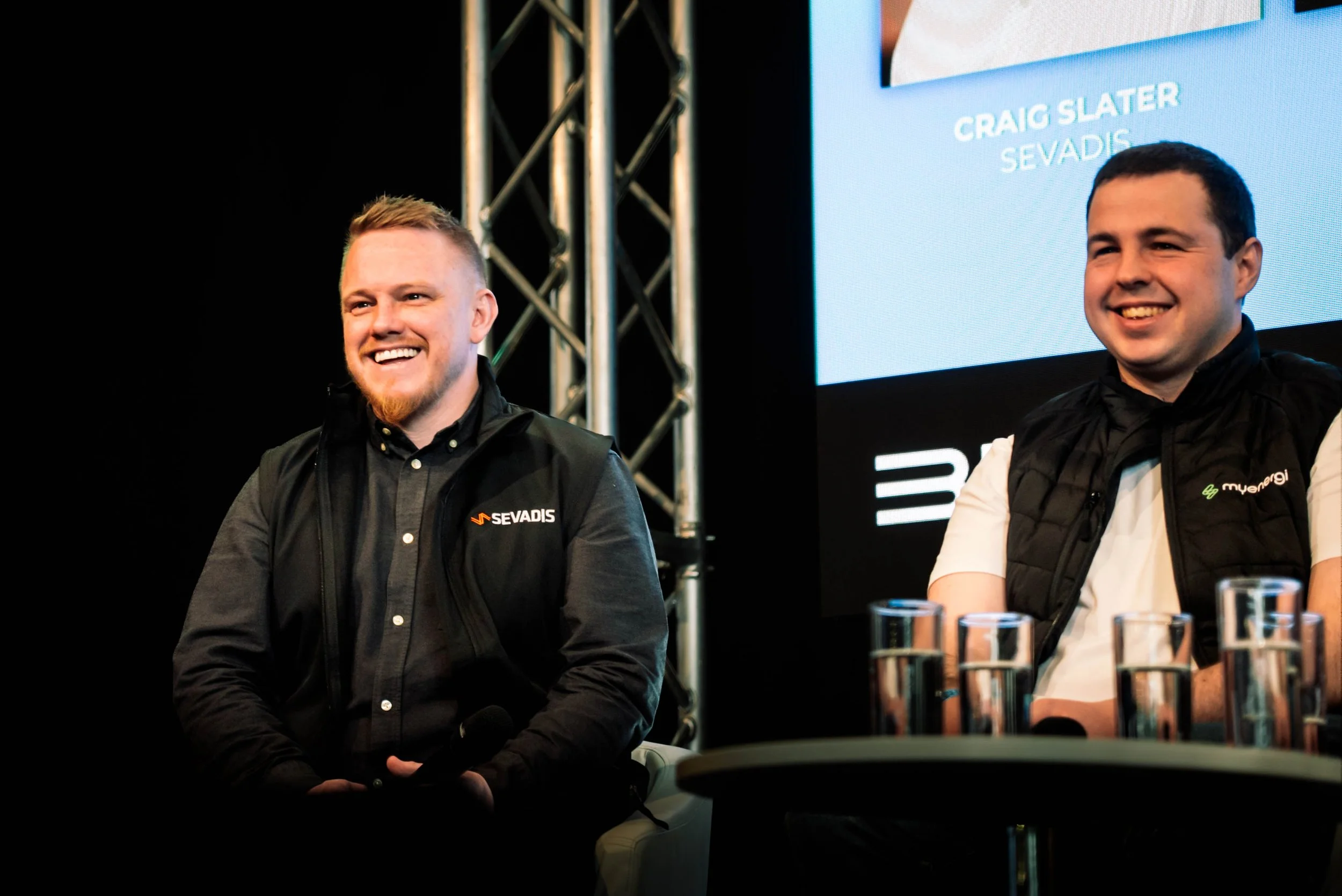 Two men sitting at a table on a stage, smiling, wearing black vests with logos, with a screen behind them displaying their names and titles, and a metallic framework to the side.