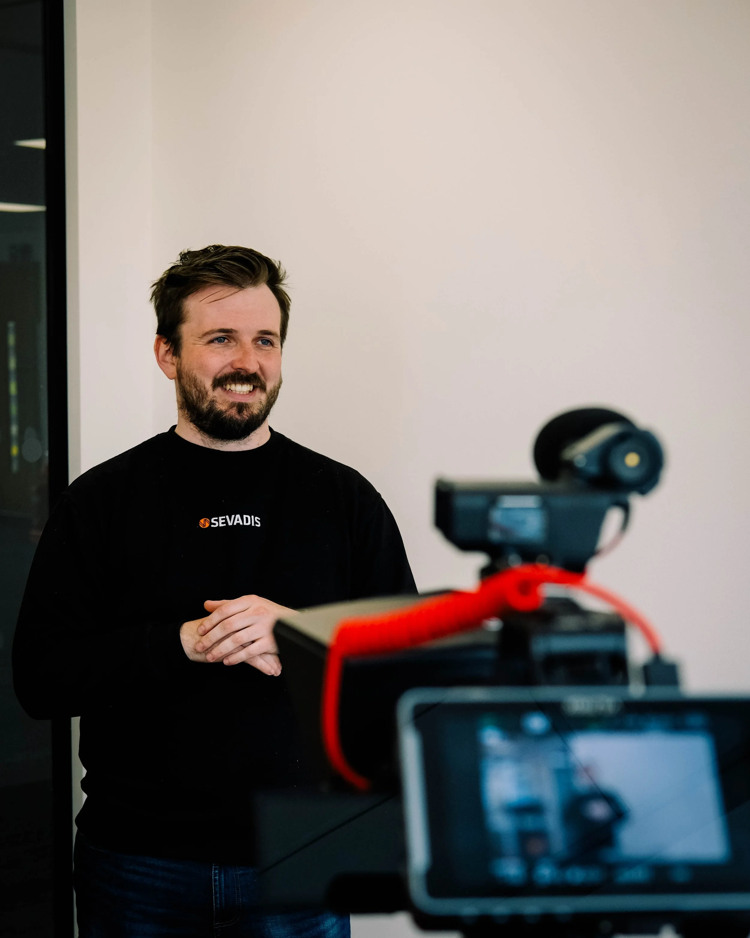 A man with dark hair and a beard smiling in front of a plain white wall, wearing a black shirt with a logo, during a recording session with a camera and microphone visible in the foreground.