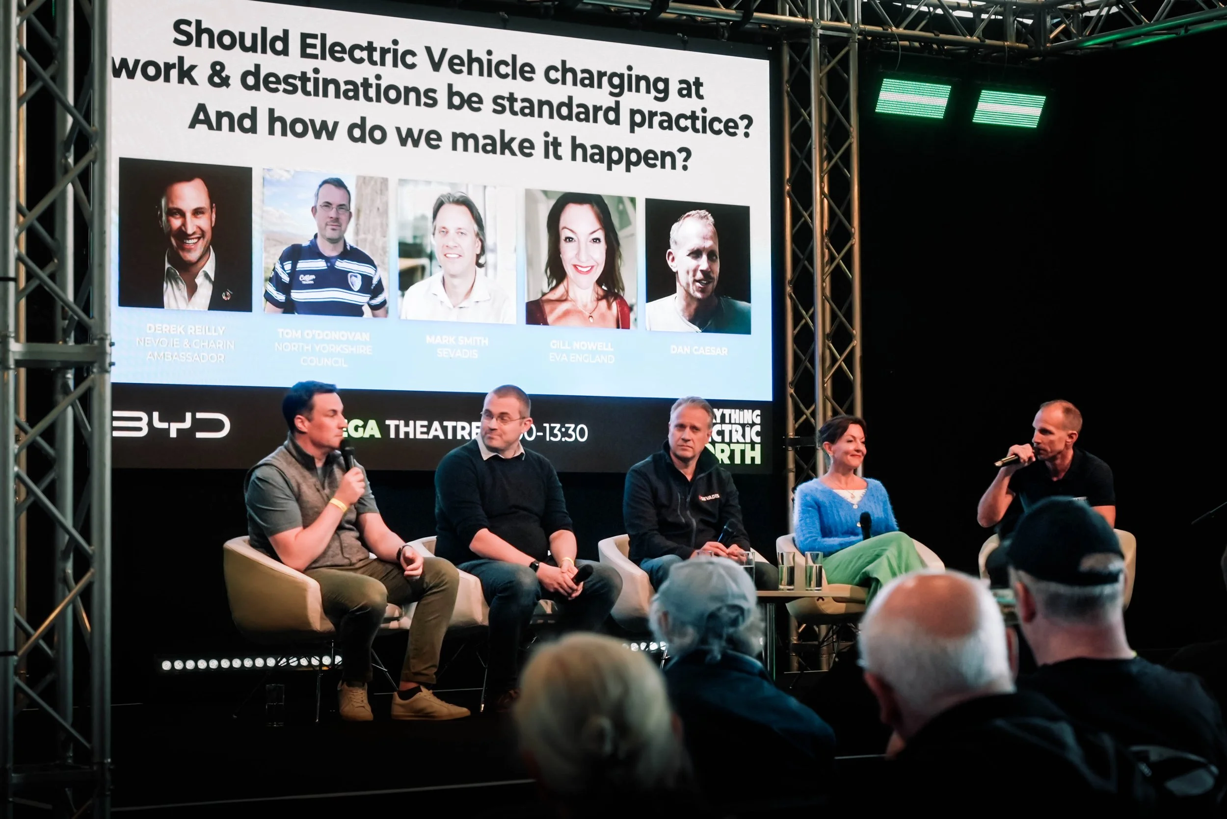 Panel discussion at an event about electric vehicle charging, featuring five panelists seated on stage with a large screen behind them displaying their photos and names.