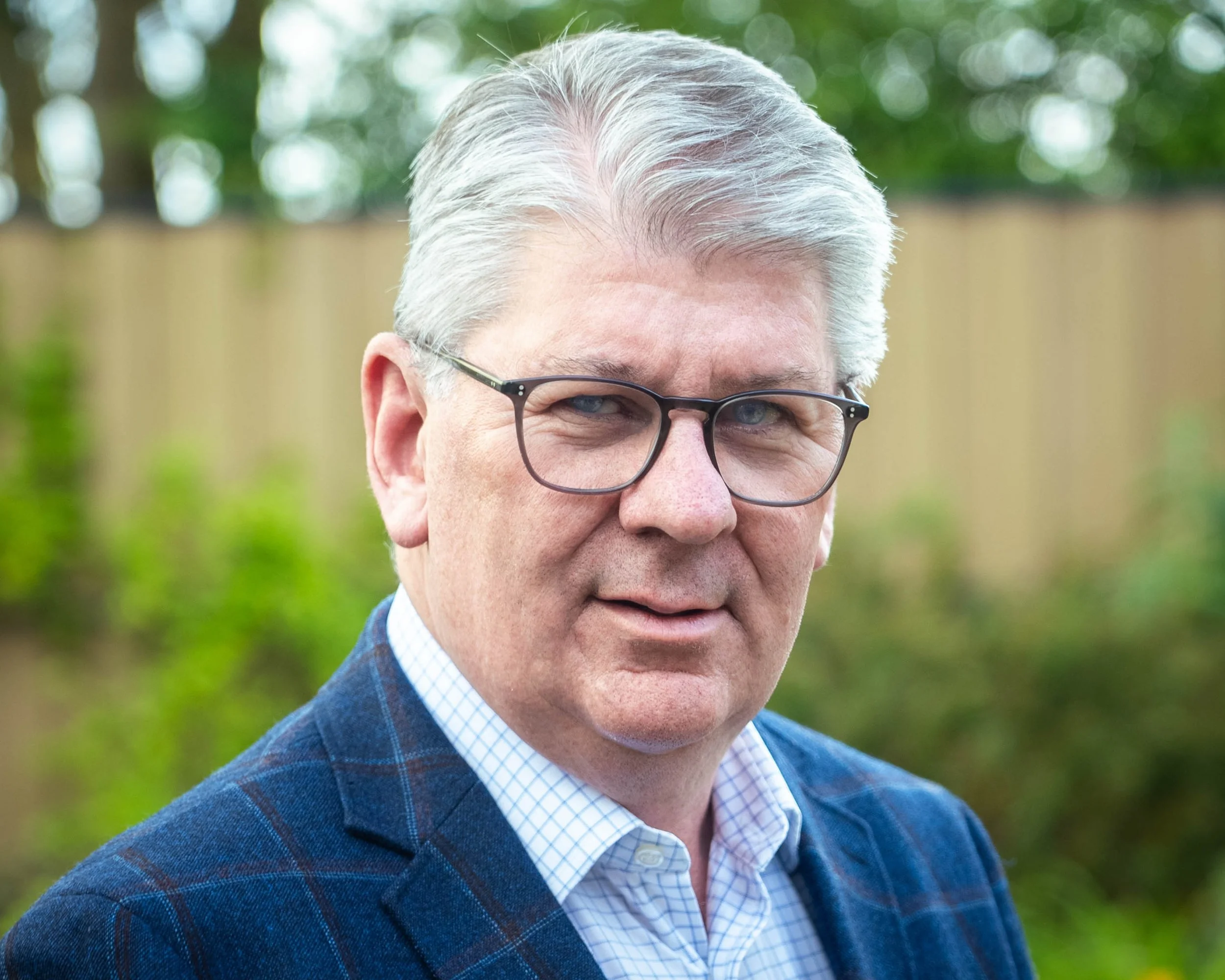 Close-up of a man with gray hair wearing glasses, a blue checkered shirt, and a dark blue plaid blazer outdoors with a green, blurred background.