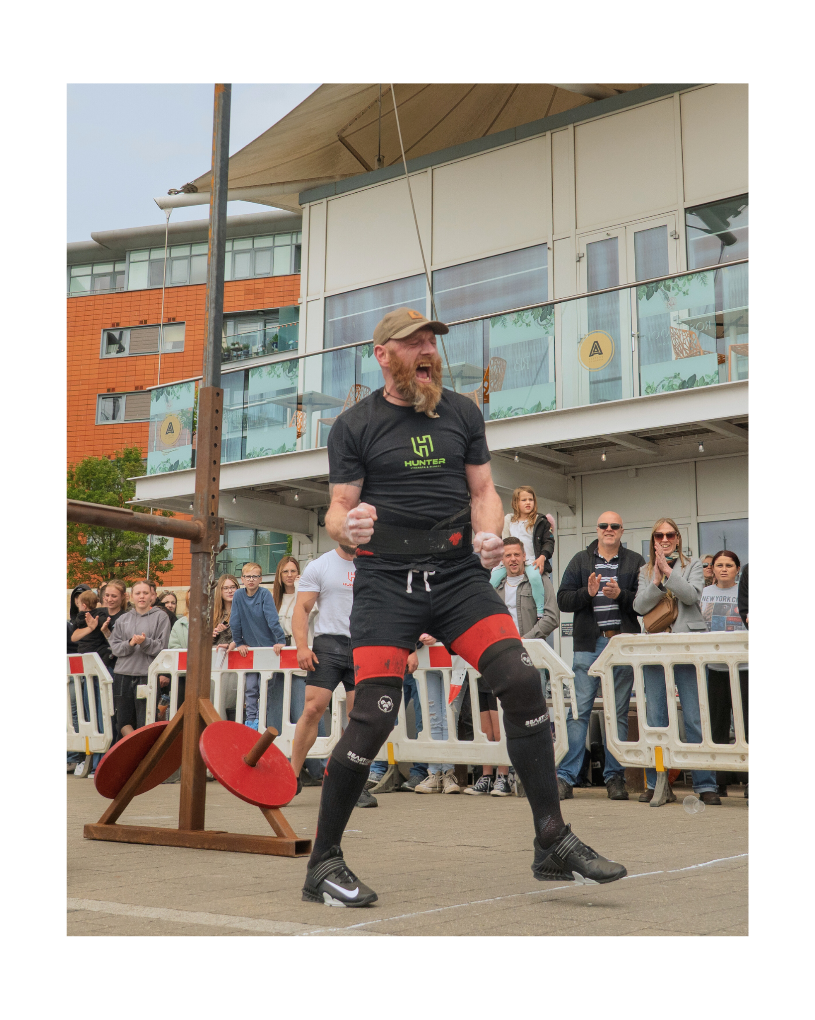 A muscular man with a beard, wearing a cap, black shorts, and sports shoes, is celebrating with clenched fists during a strongman competition, with an audience watching behind a white safety barrier and a vintage car on display in the background.