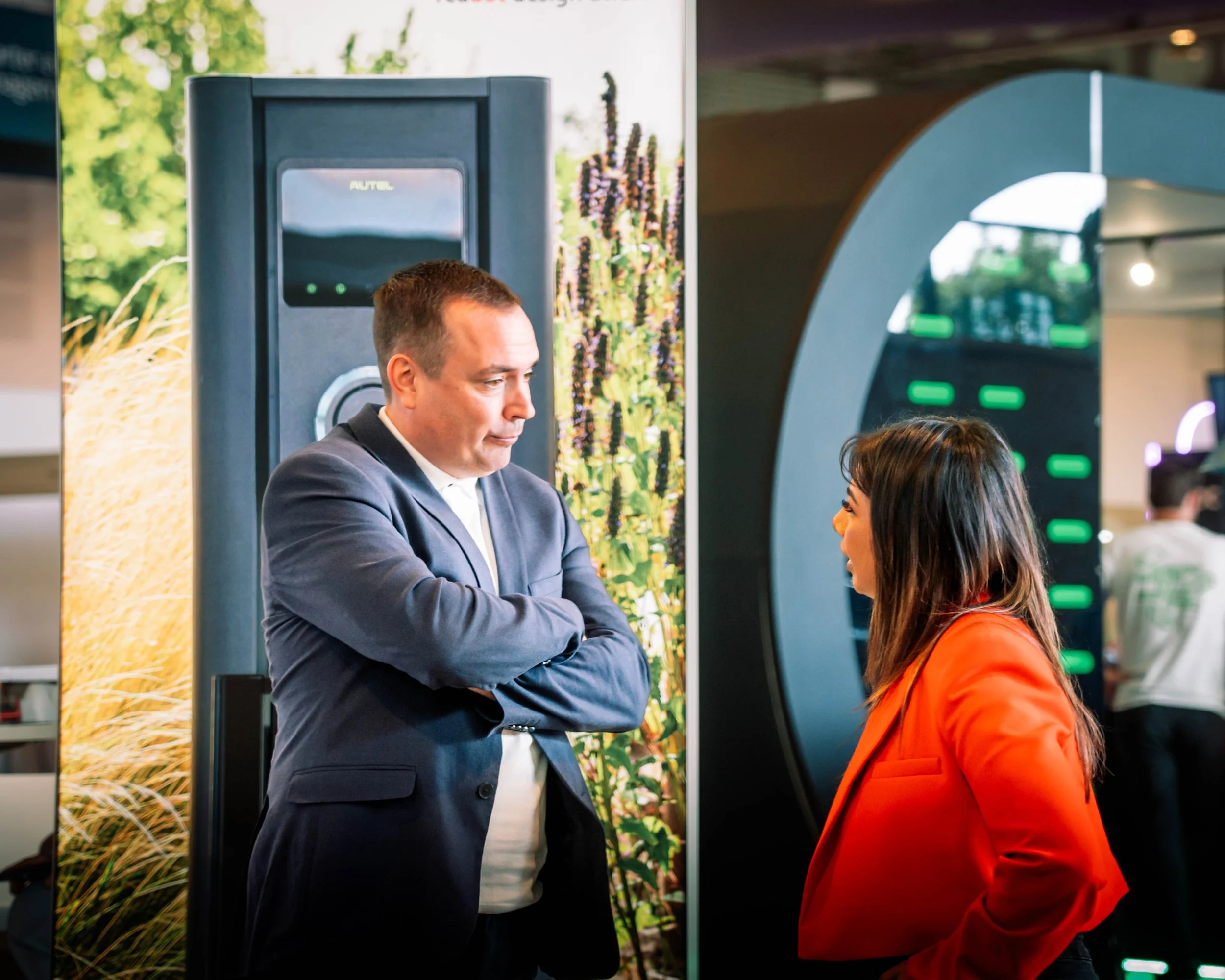 A man in a suit with short hair and a woman with black hair wearing an orange jacket are having a conversation in a modern indoor space with digital screens and nature-themed decor.