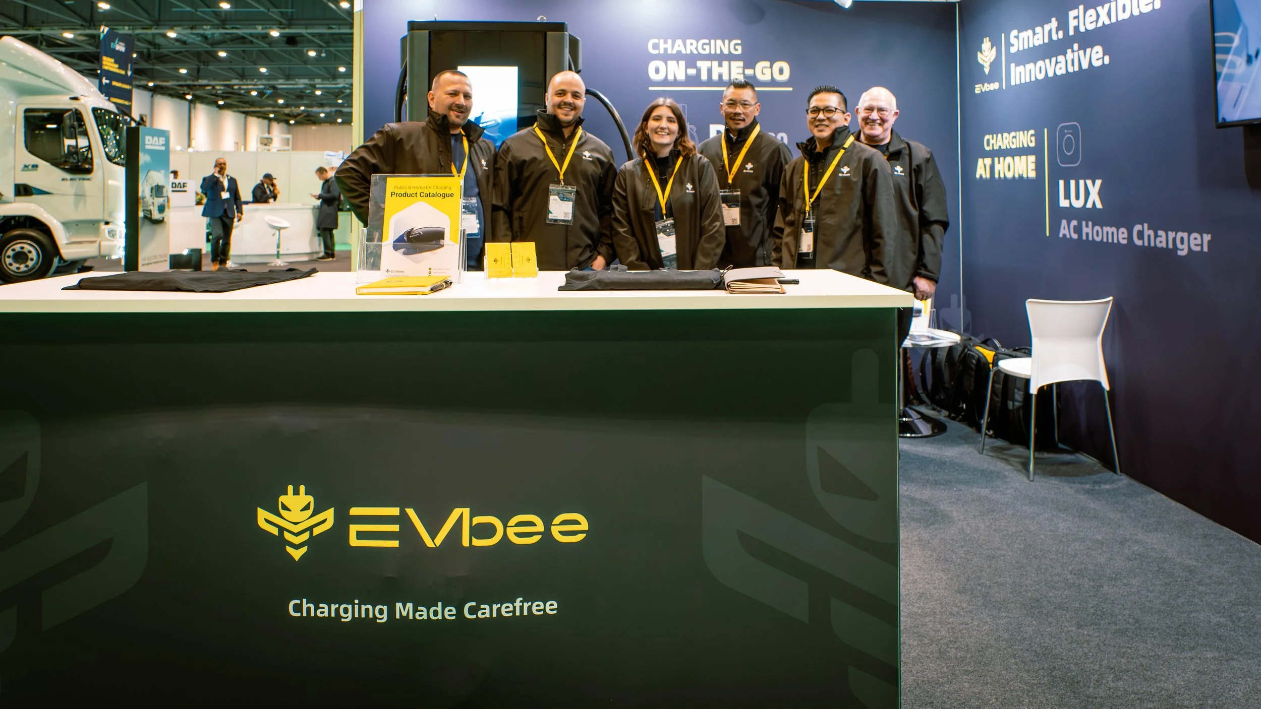 Group of six people standing behind a booth table at an exhibition, showcasing EVbee electric vehicle charger products, with banners displaying EVbee branding and features.