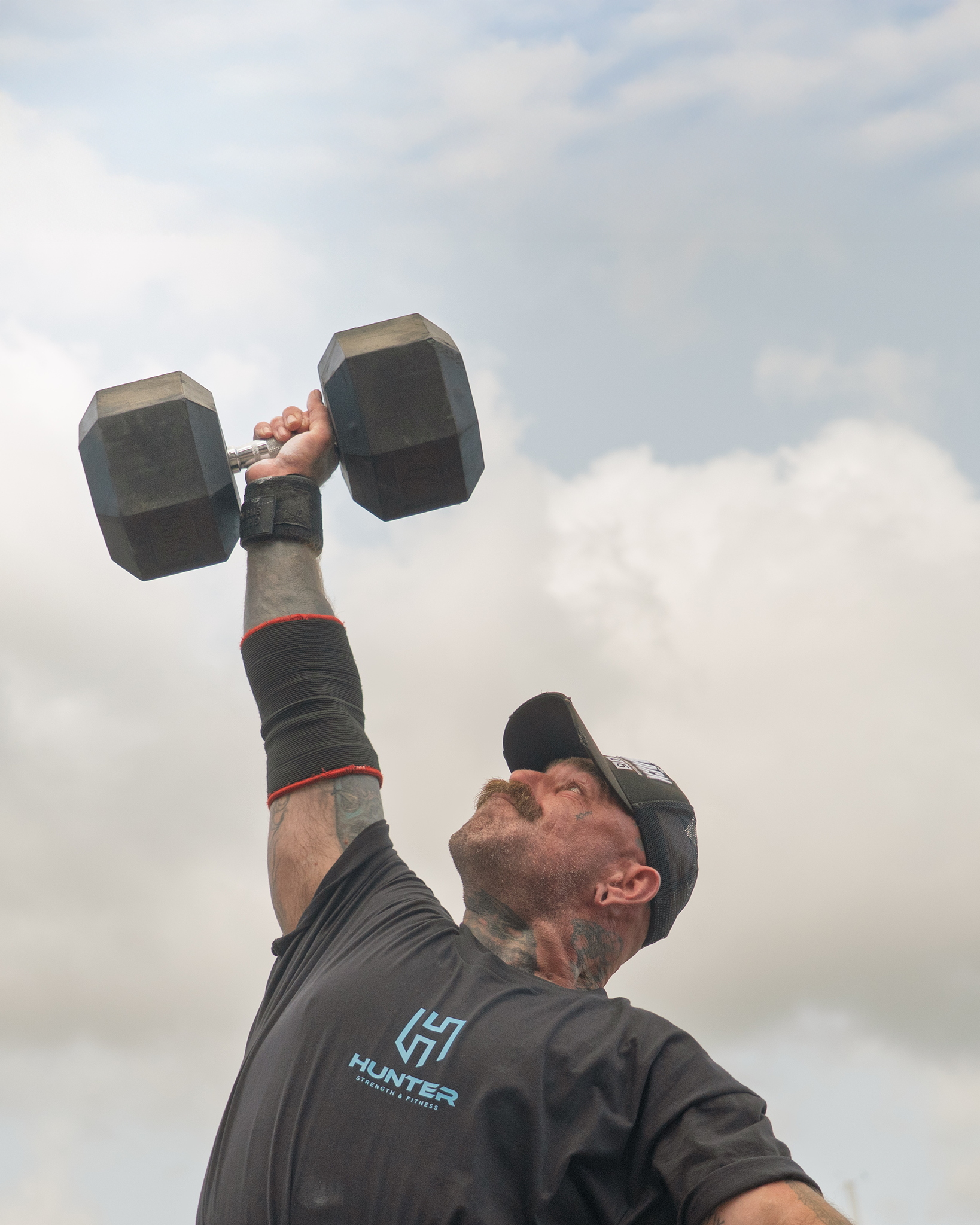 A man wearing a black cap and black shirt with a logo, exercising outdoors and lifting a dumbbell over his head, with a partly cloudy sky in the background.