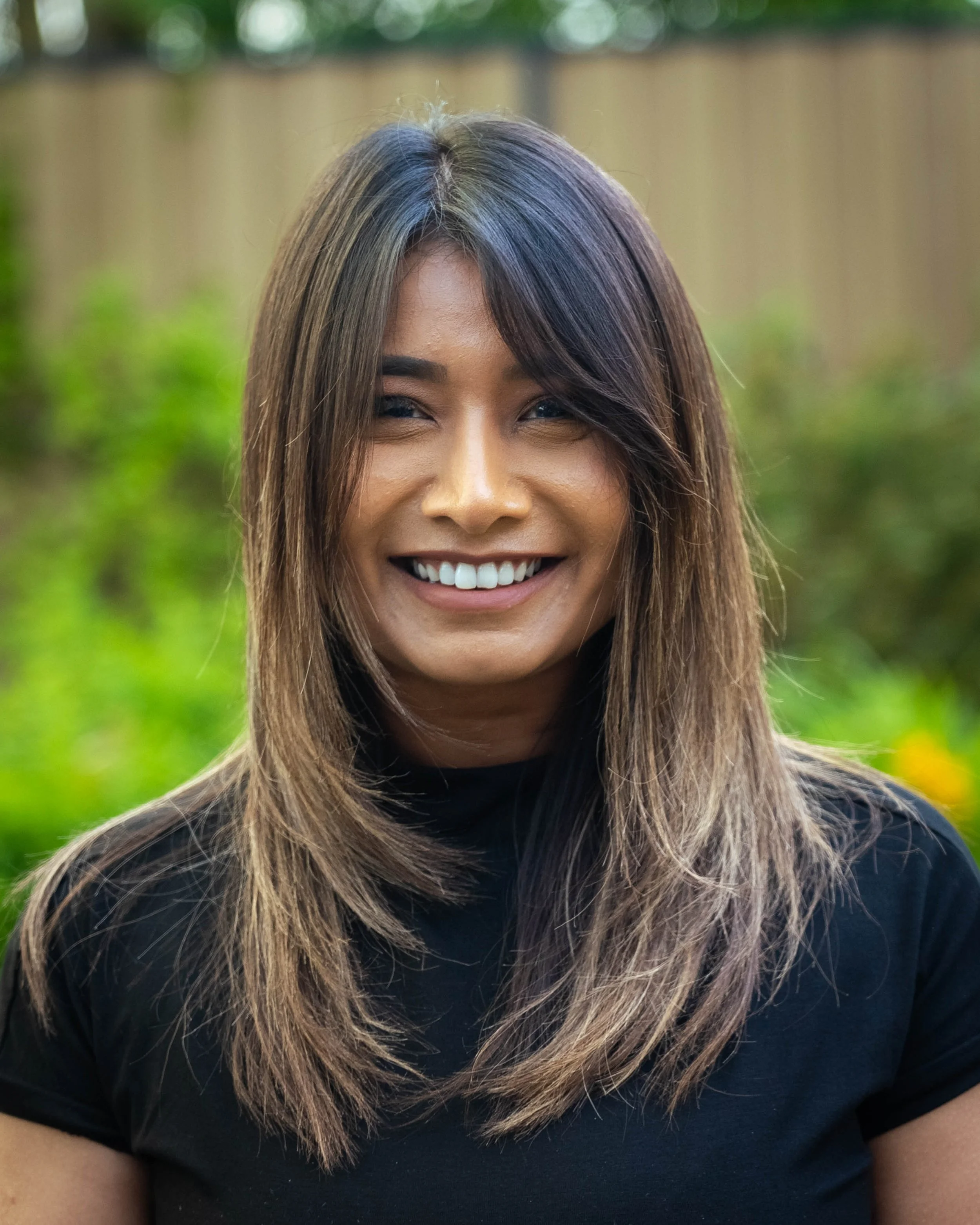 Close-up of a person with long, brown layered hair, smiling, wearing a black top, outdoors with blurred green and wooden background.