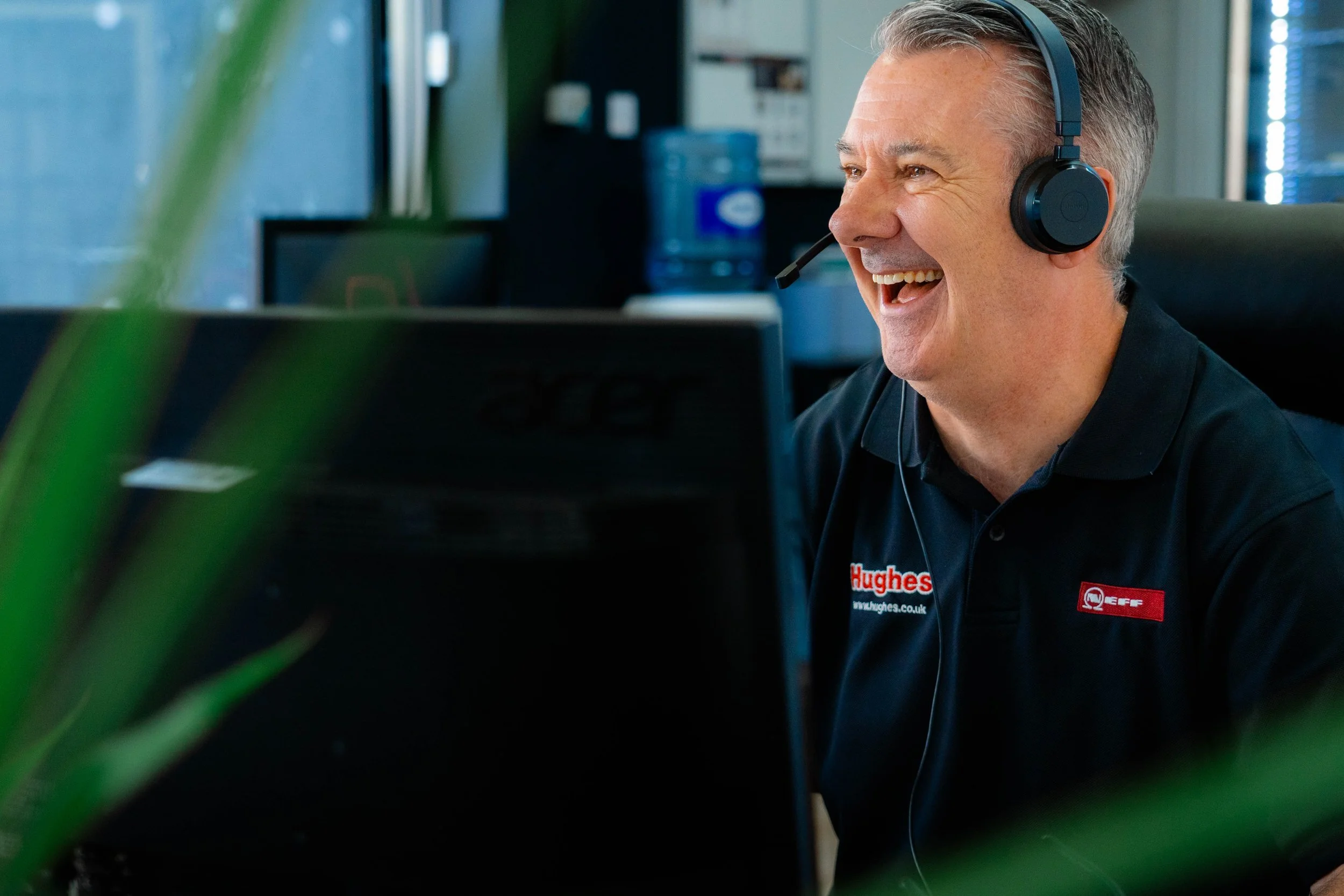 A middle-aged man with gray hair wearing a black Hughess branded polo shirt, a headset with microphone, smiling and working at his computer in an office setting.