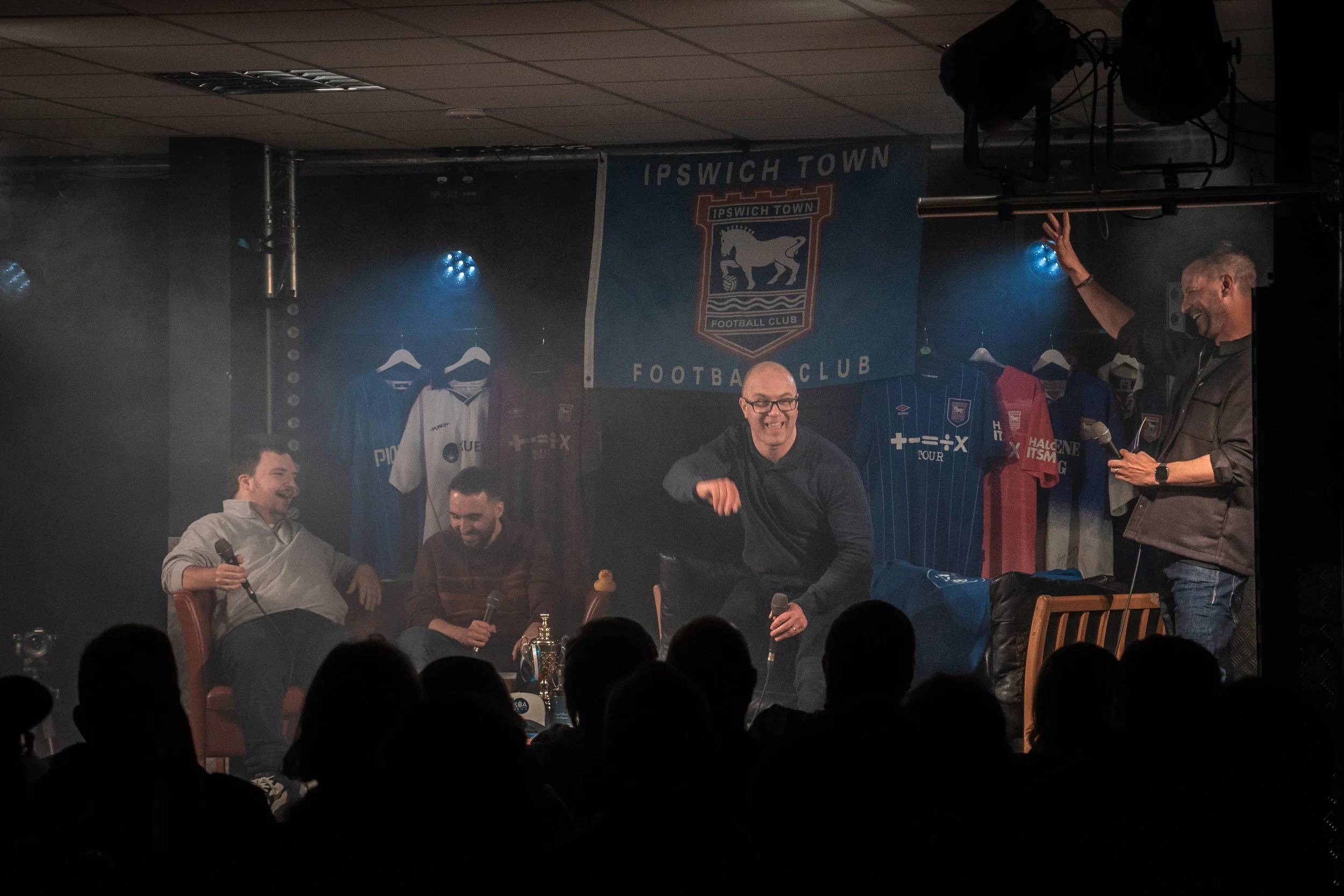 A group of people sitting on stage with microphones, soccer jerseys hanging in the background, and a Ipswich Town Football Club banner hanging on the wall behind them.
