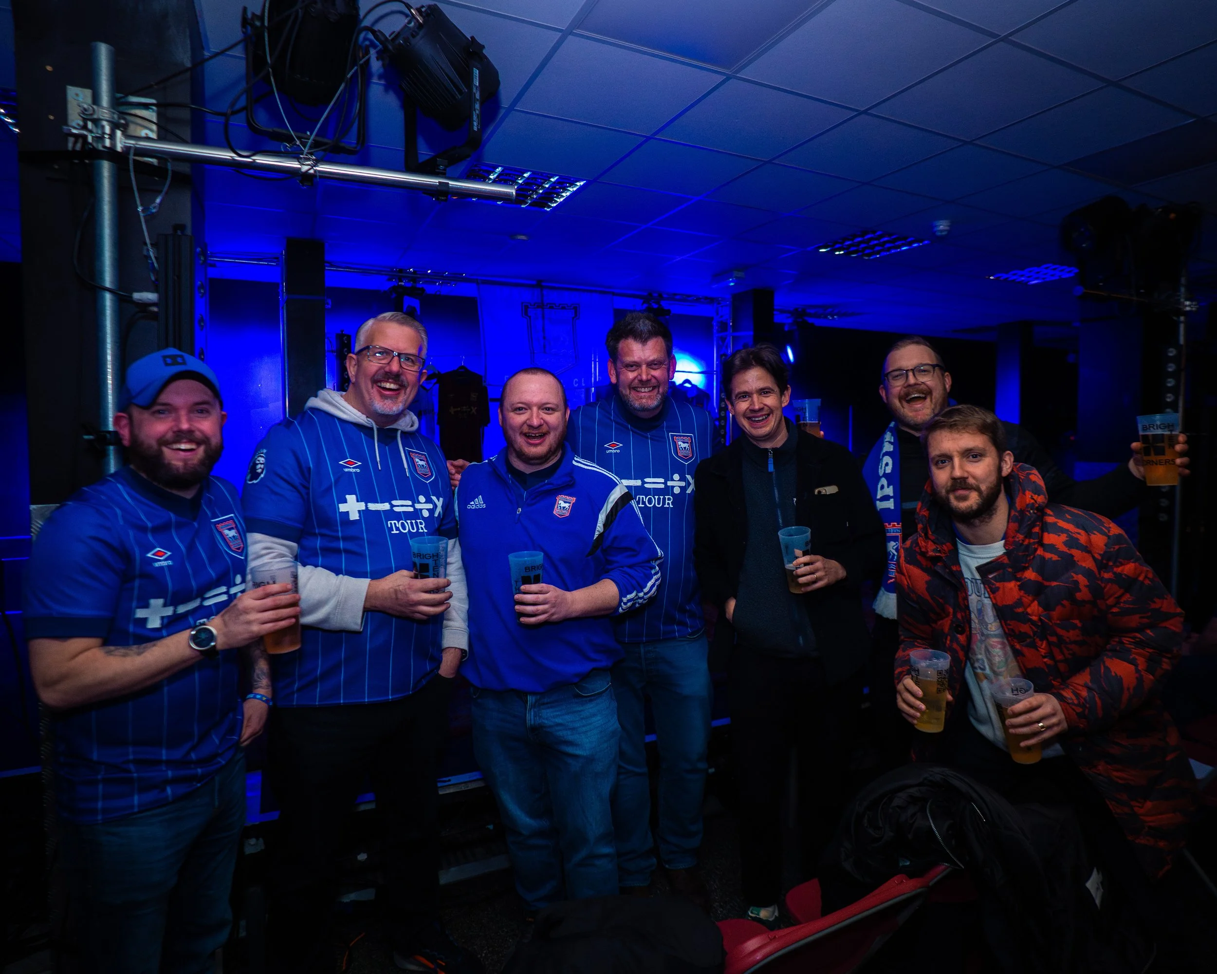 A group of seven men smiling and holding drinks at an indoor event, wearing sports jerseys and casual clothing, with blue lighting and equipment in the background.