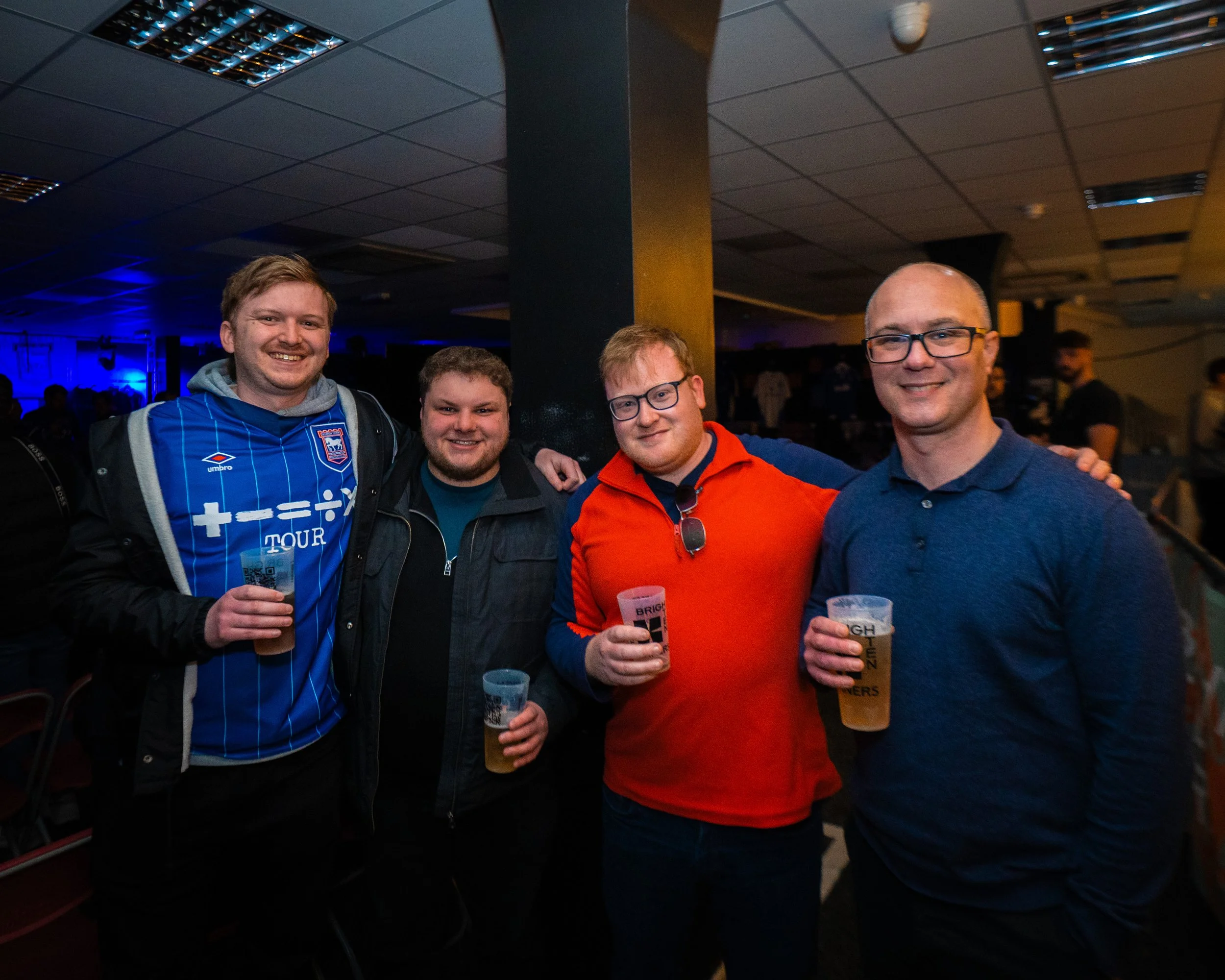 Four smiling men standing closely together in a bar or pub, holding drinks, with a dark ceiling and some background patrons.