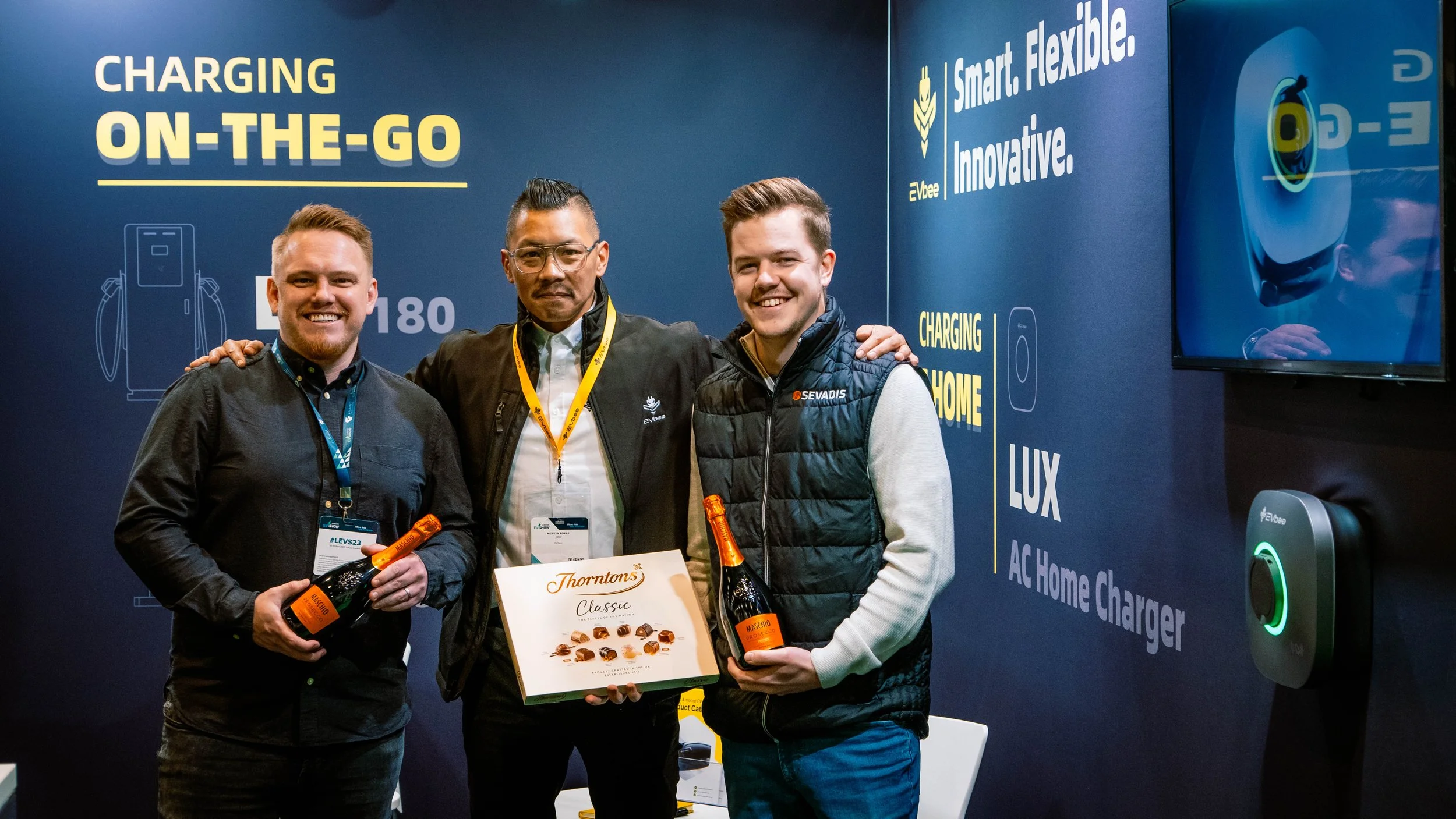 Three men standing together at an event, holding bottles of sparkling wine, in front of a blue backdrop with text about smart, flexible, innovative charging solutions, and an electric vehicle charger on the right side.