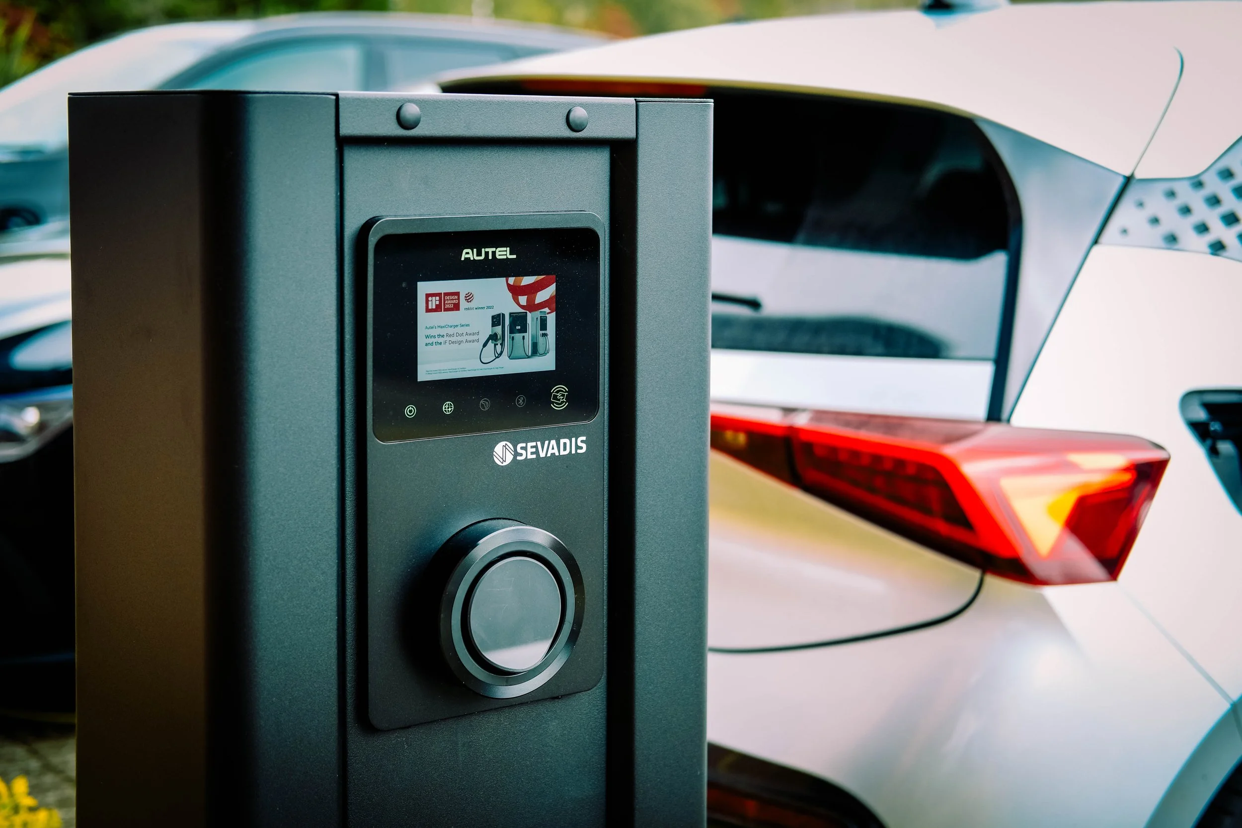 Electric vehicle charging station with a digital display, branded Autel, in front of a white electric car with a red tail light.