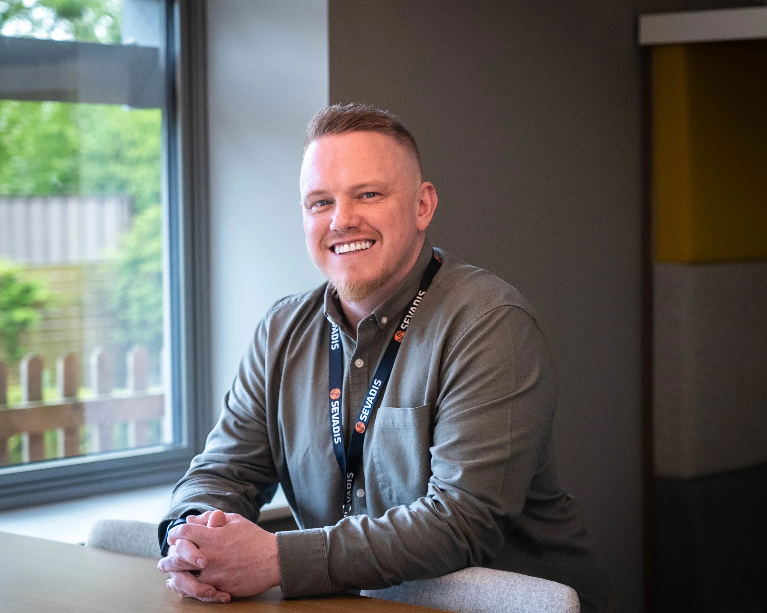 A smiling man with short brown hair and a beard, wearing a gray button-up shirt with a black lanyard, seated at a table in a room with a large window showing a backyard with trees and a wooden fence.