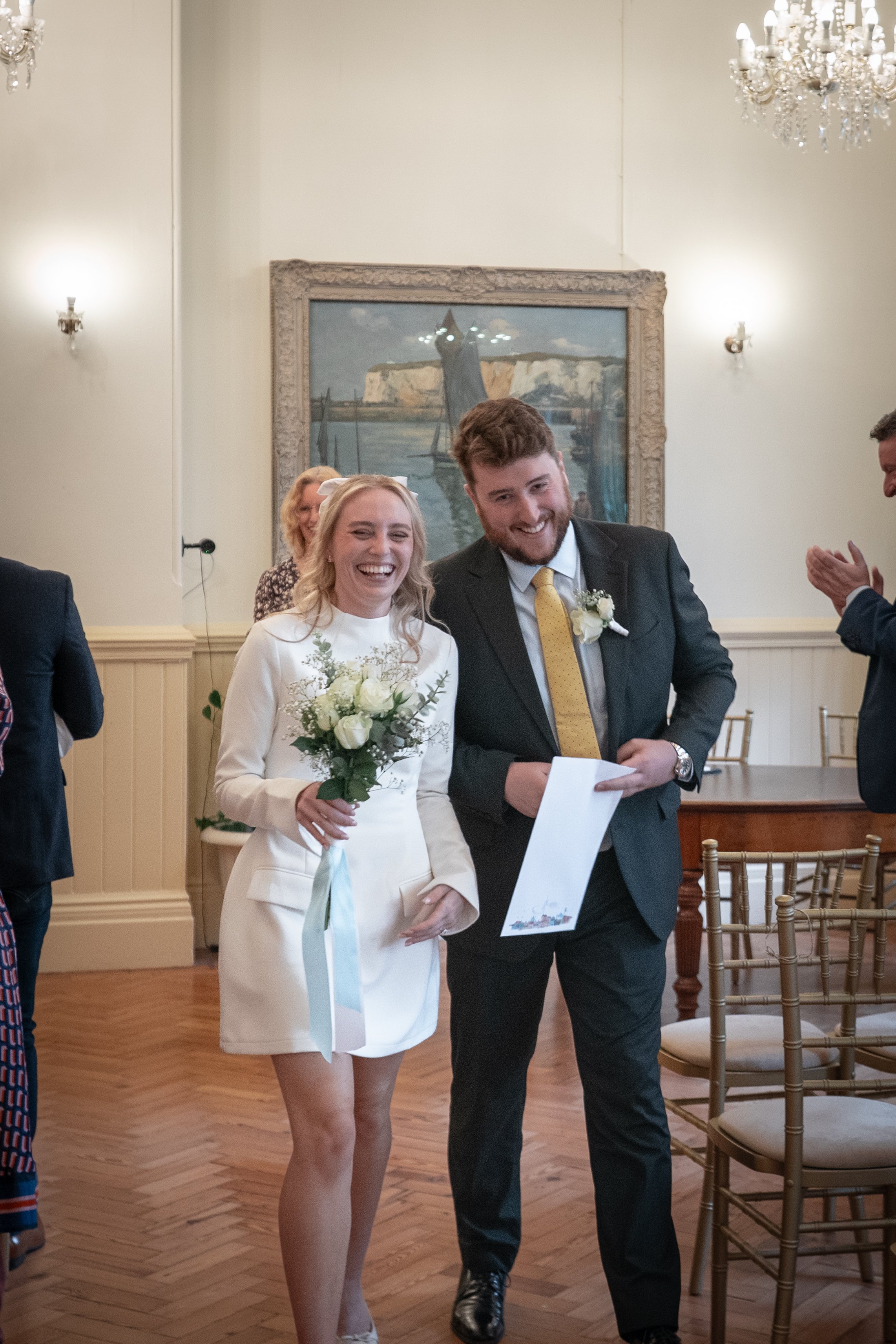 A smiling bride in a white dress with a bouquet in her hand standing next to a smiling groom in a suit and yellow tie. They are at a wedding ceremony, with chairs and people clapping around them in an elegant room with chandeliers and a framed painti