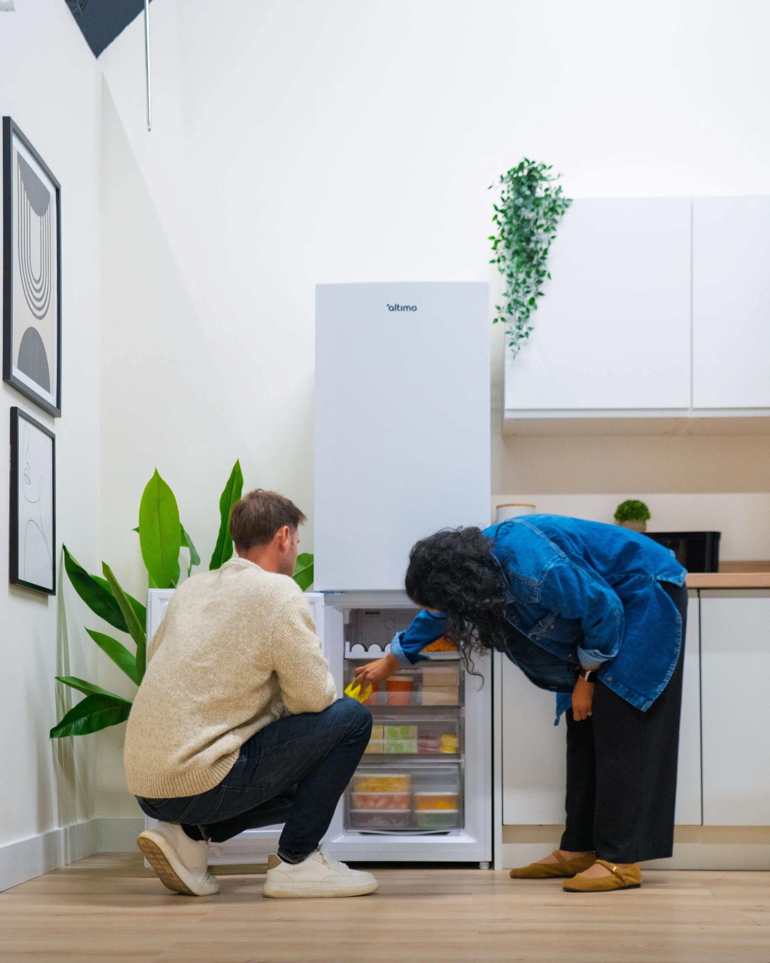 Two people, one kneeling and one bending, are looking at a small appliance or container on a low shelf in a white kitchen or living space, with green plants and framed artwork on the wall.