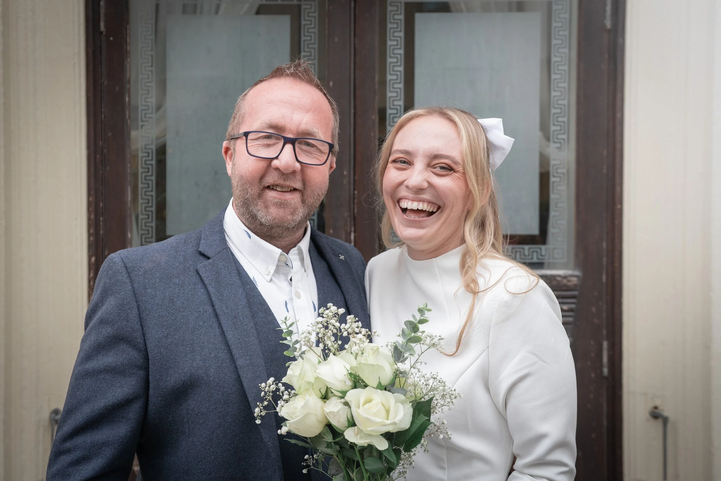 A happy bride and groom standing outside a building, the groom wearing glasses and a blue suit, and the bride with a white dress and a white bow in her hair, holding a bouquet of white roses and greenery.