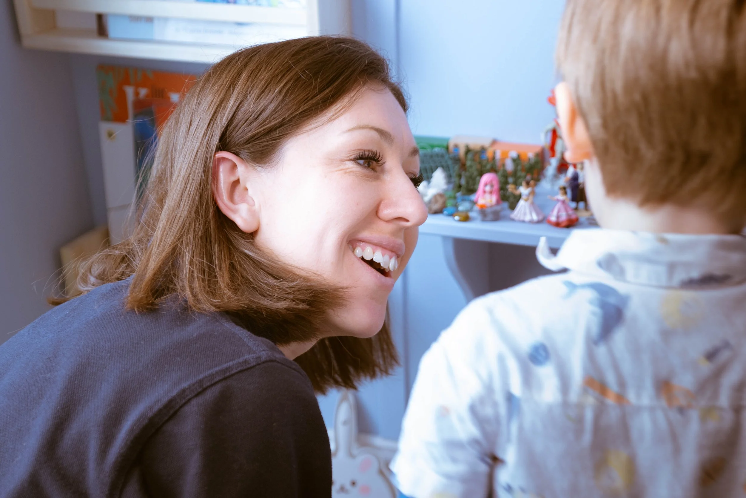 A woman with brown hair smiling and looking at a young child with brown hair facing away, with the background showing a shelf with figurines and toys.