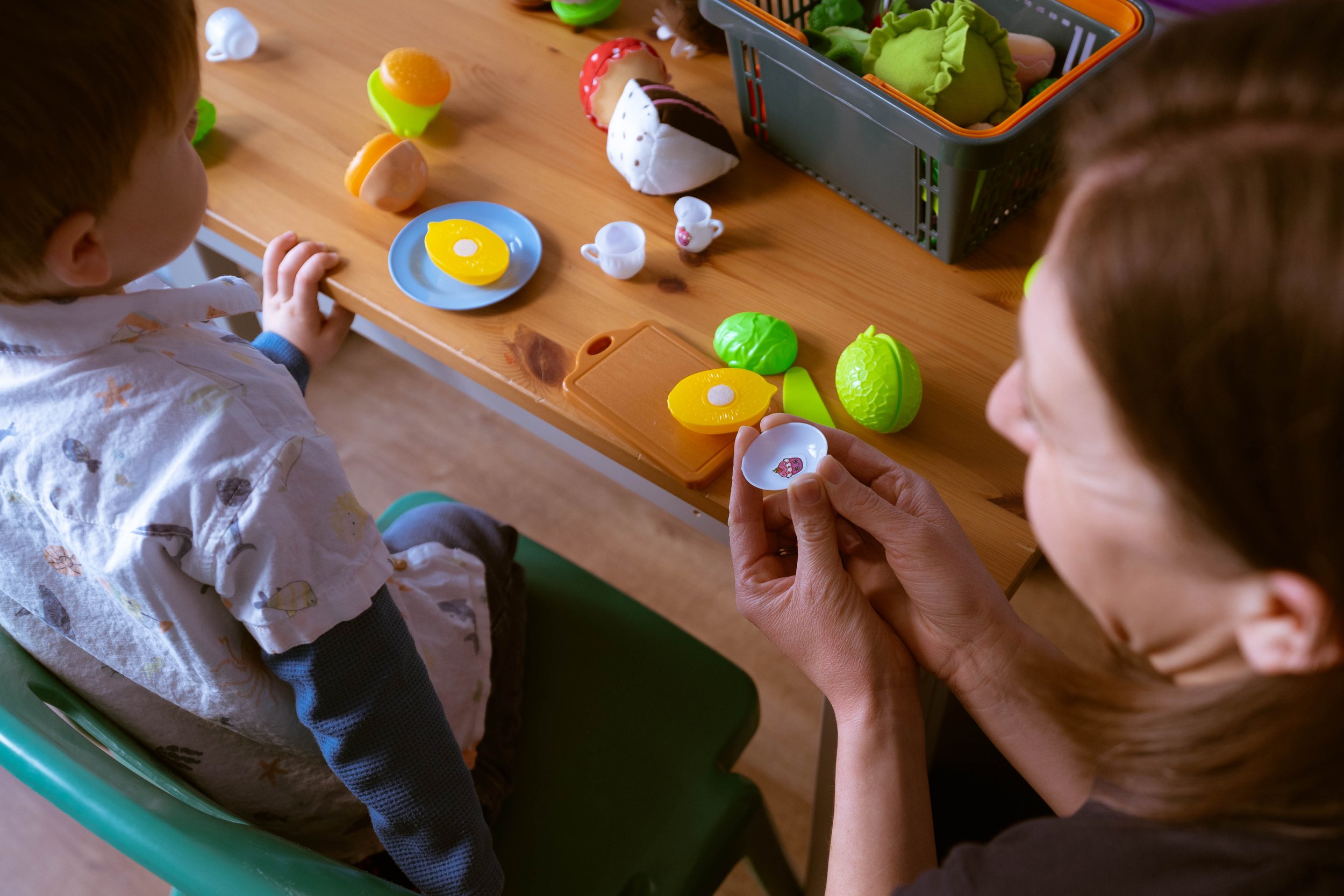 Children playing with toy food and dishes at a table with a basket of plush toys.