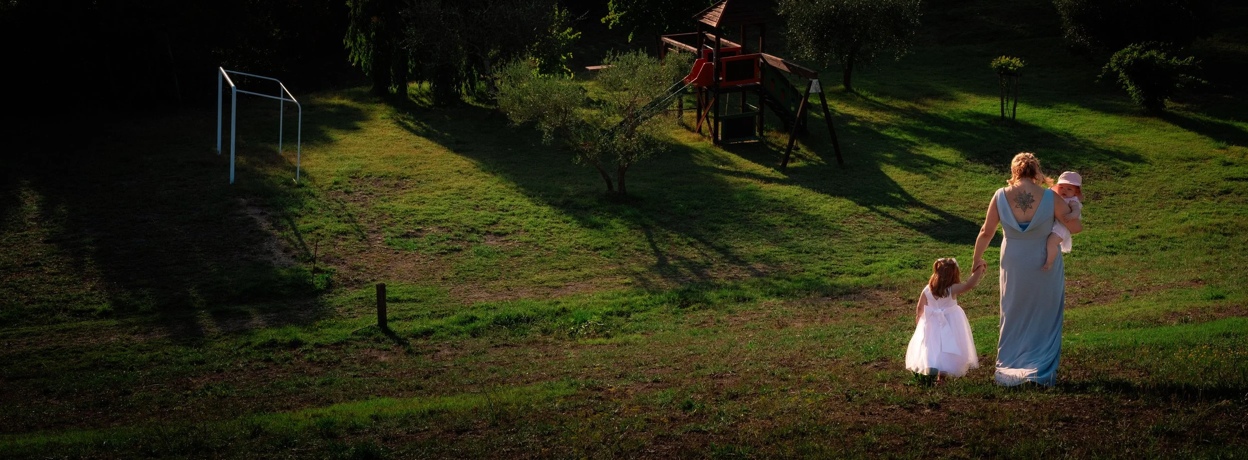 A woman with a tattoo on her back holding a baby in a white hat walking hand-in-hand with a young girl in a white dress in a grassy park at sunset, with a playground and trees in the background.