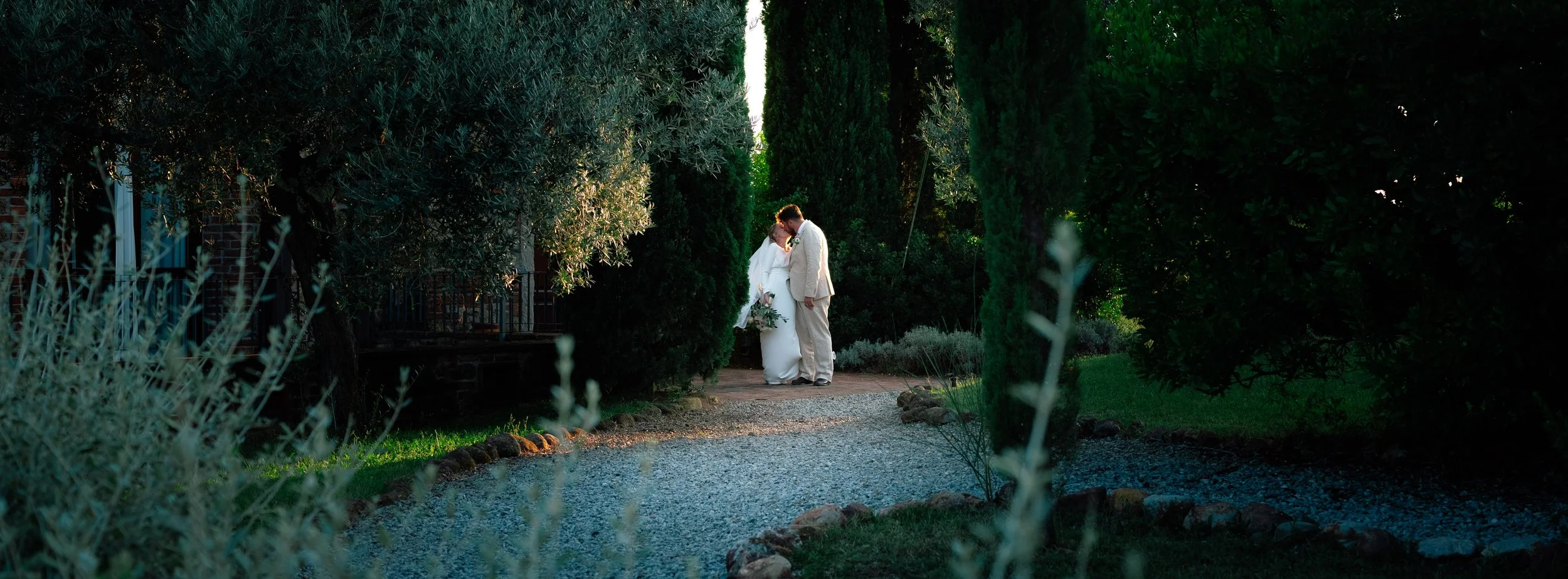 A bride and groom stand close together on a garden pathway, surrounded by tall green trees and bushes during sunset.