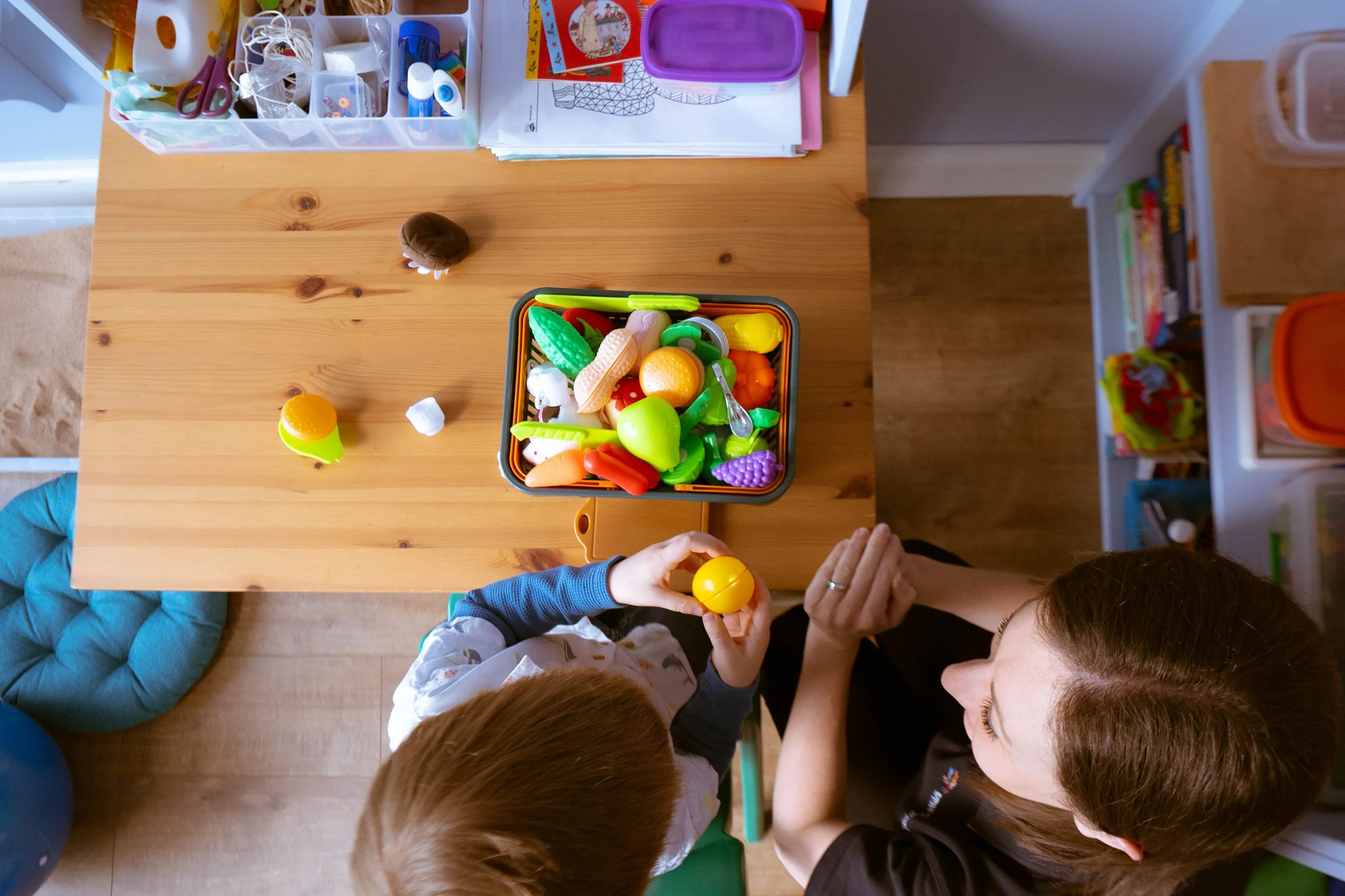 Children gathered around a wooden table playing with colorful plastic toy food and utensils. The table has a container filled with various toy foods, and one child is holding a yellow toy egg. There are storage drawers, books, and craft supplies in t