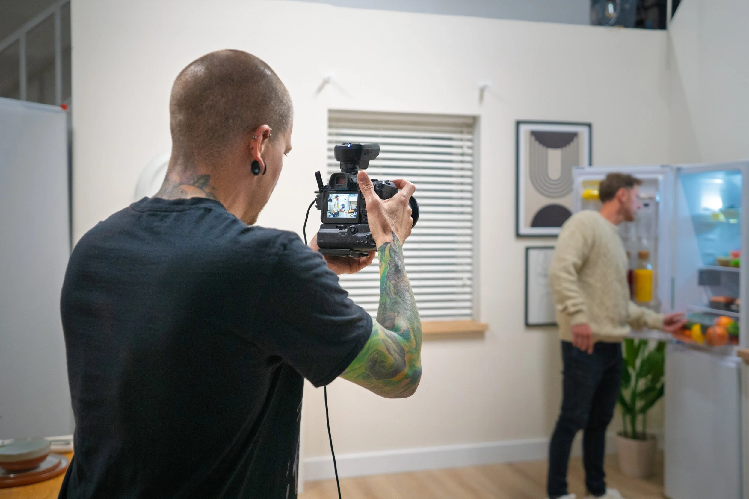 Person with tattoos on arms and multiple ear piercings taking a photograph in a modern, minimalistic interior space with another person in the background near a fridge with open door.