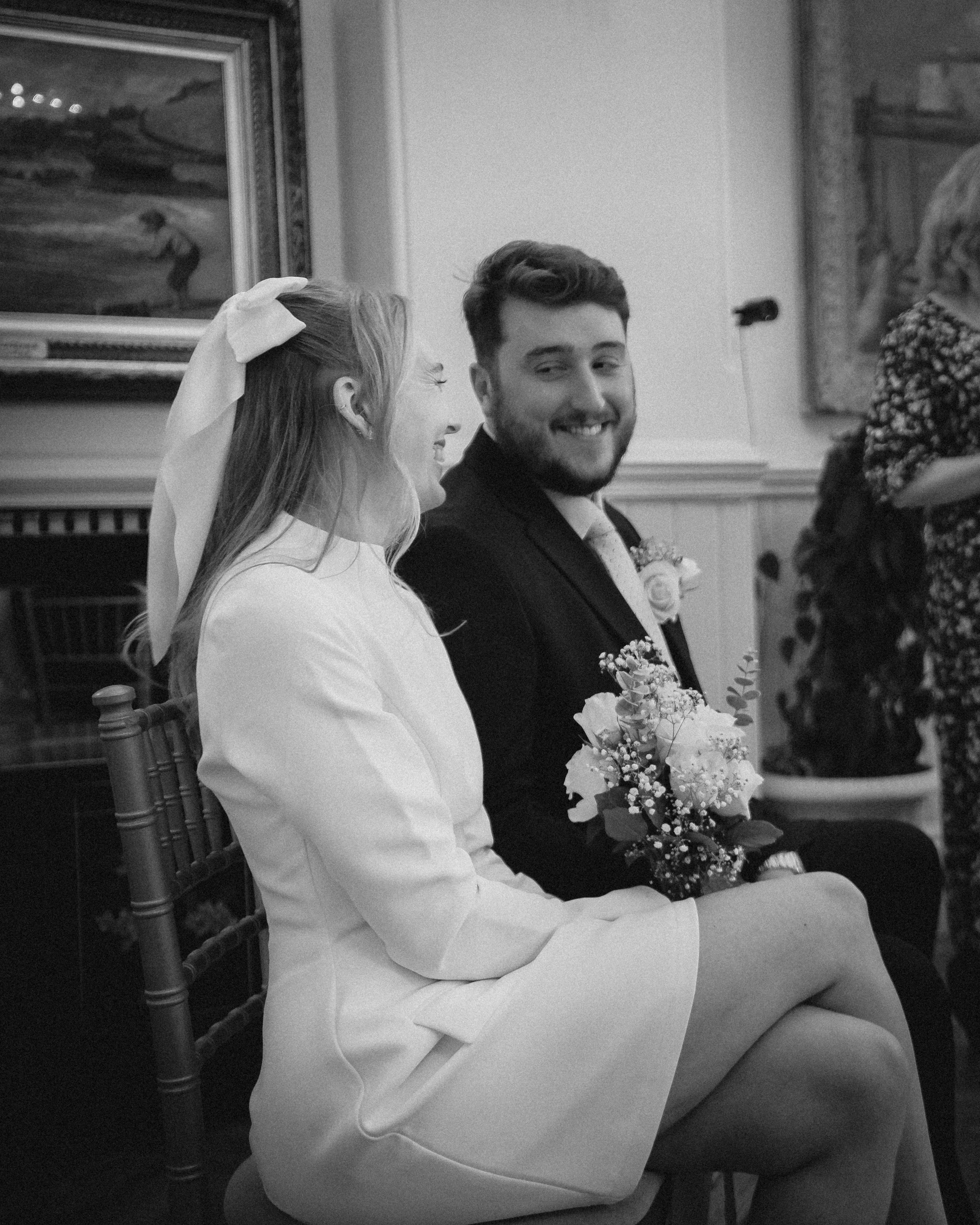 A bride and groom sitting side by side, dressed in wedding attire in a room with paintings and a microphone in the background.