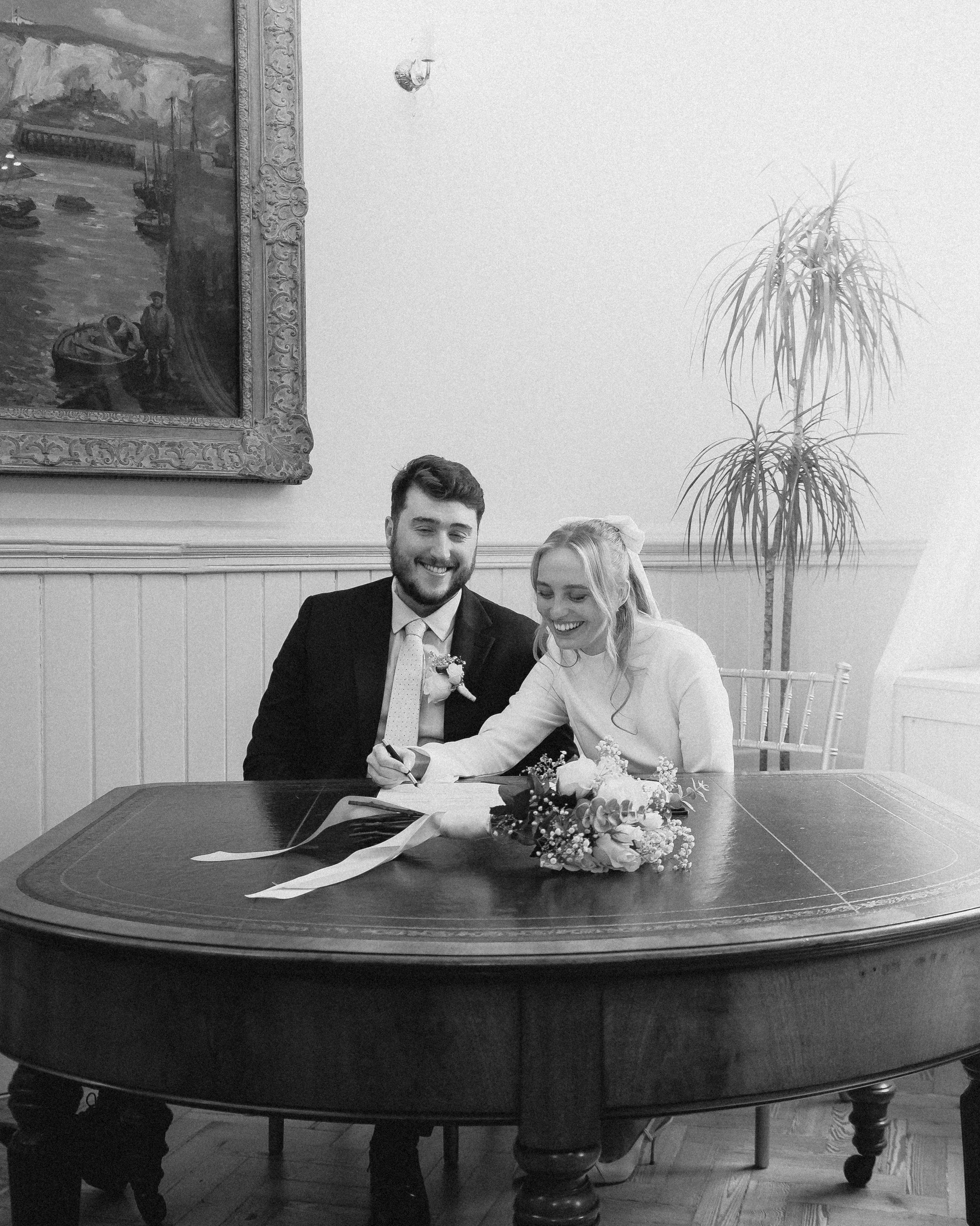 A smiling man and woman, dressed in wedding attire, sit together at a table signing documents, with a bouquet of flowers in front of them. Behind them, a potted plant stands next to framed artwork on a white wall.