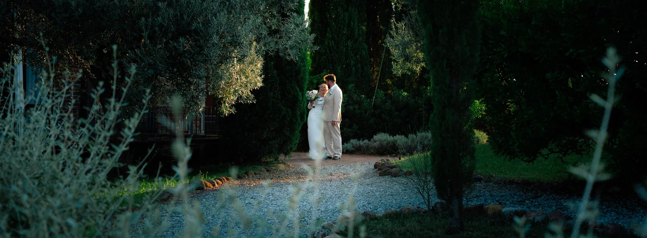 A bride and groom standing together in a lush garden, celebrating during sunset with greenery and trees surrounding them.