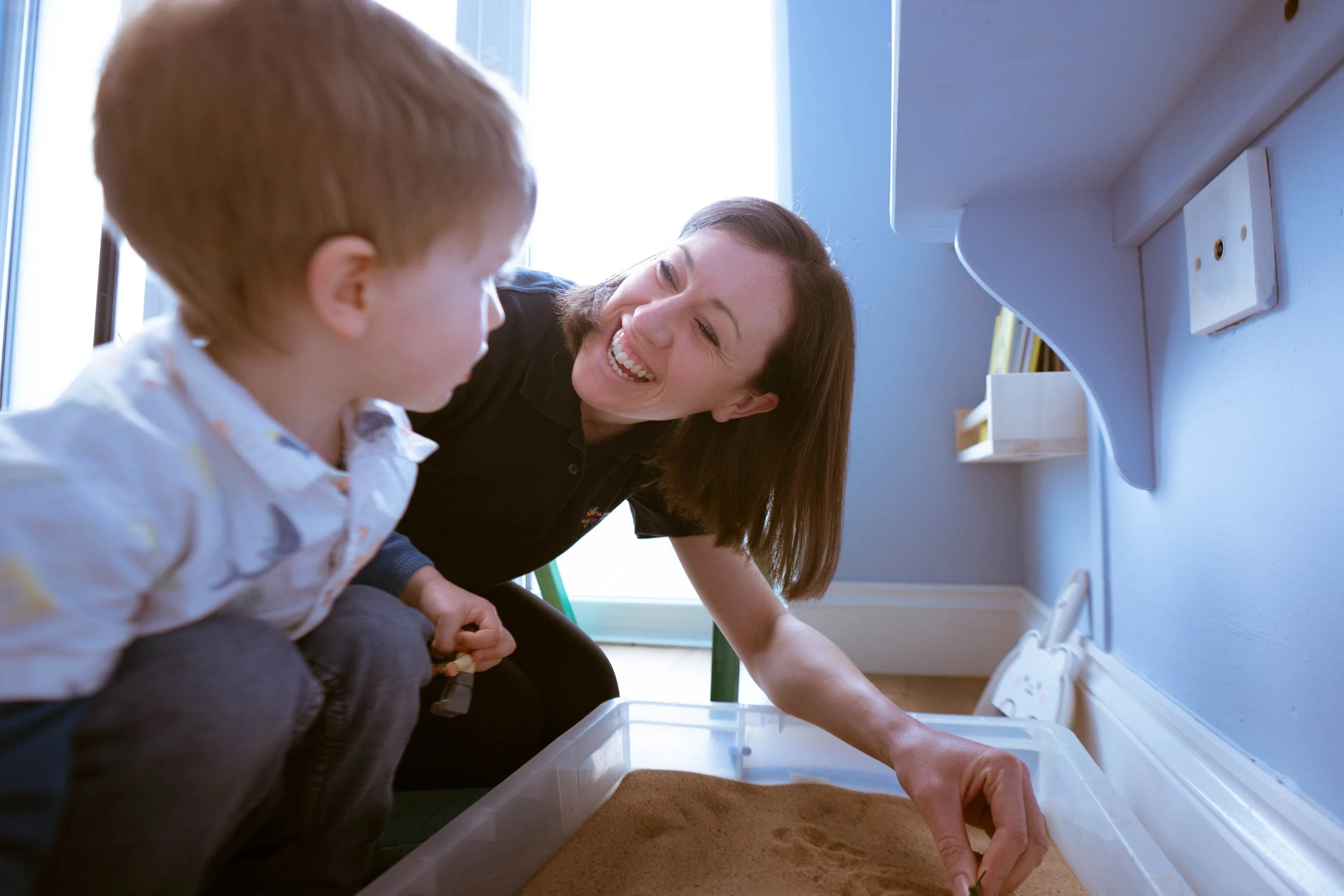 A woman and a young boy sitting by a window, smiling and engaging in an activity involving a plastic container and brown granular material, possibly sand or dirt, inside a well-lit room.