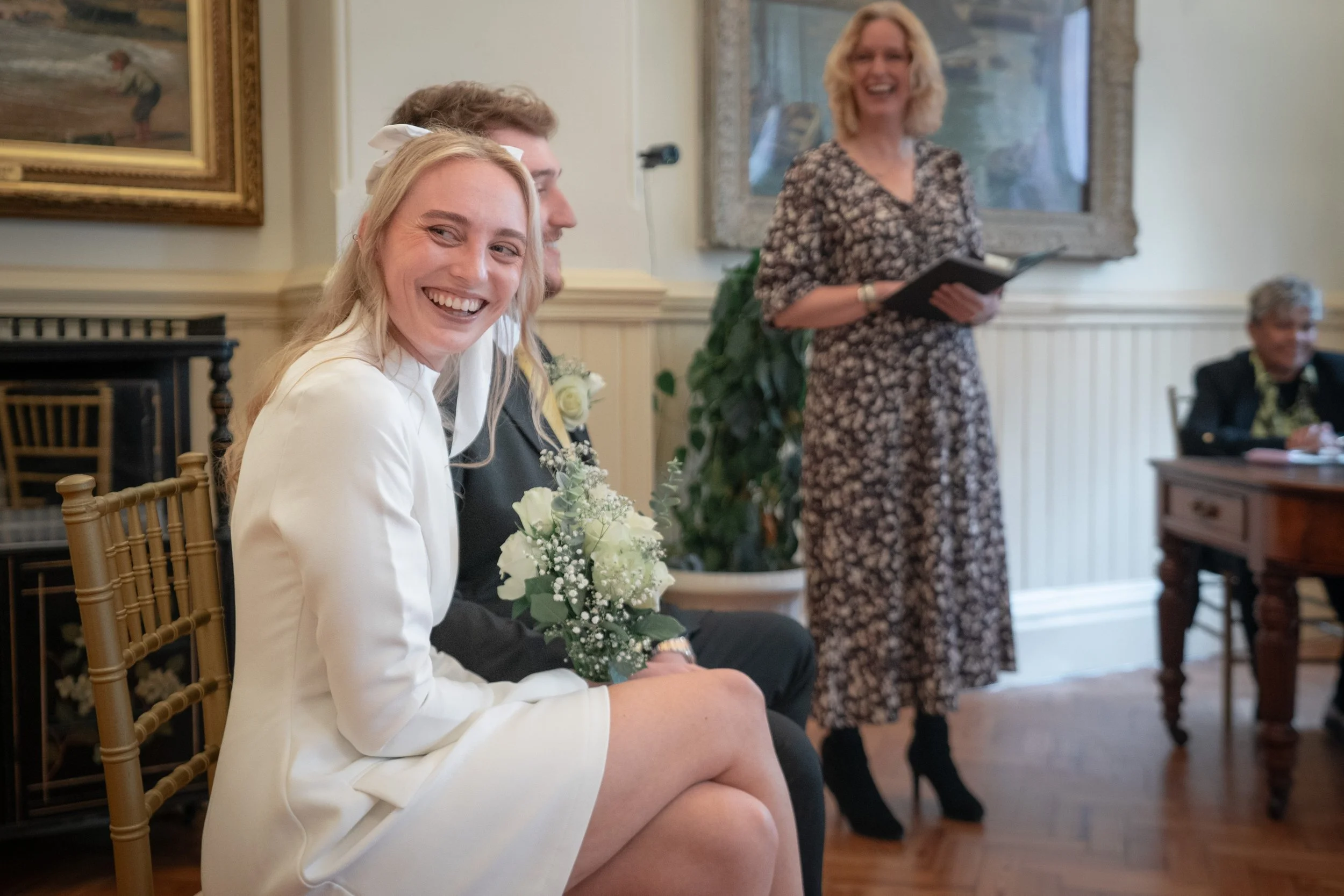 A woman in a white dress smiling, sitting with a bouquet of white flowers, in a room with framed artwork on the walls. A woman in a patterned dress is standing and speaking, holding a folder, with other people seated in the background.