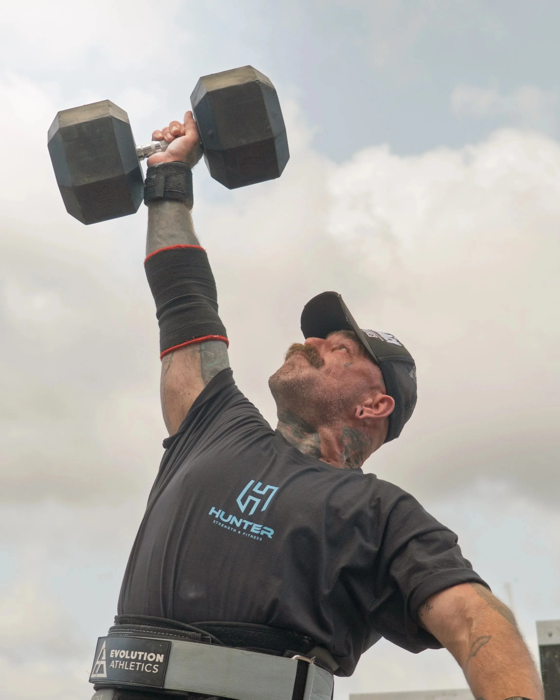 A man lifting a dumbbell during an outdoor workout, wearing a black t-shirt, a sports cap, and a weightlifting belt, with a cloudy sky in the background.