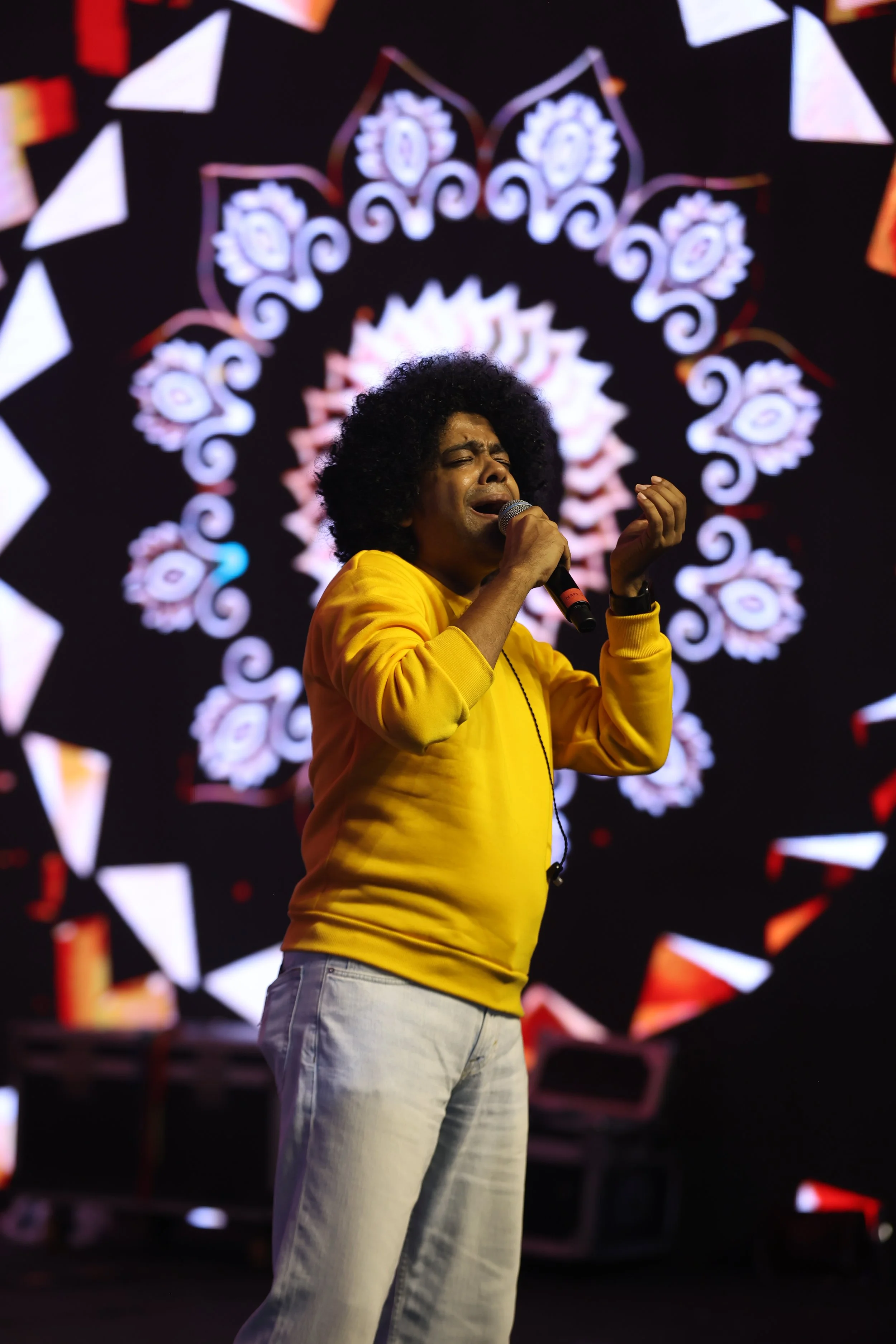 A man with curly black hair singing into a microphone on a stage with a colorful, circular, floral pattern in the background, wearing a yellow hoodie and light-colored pants.