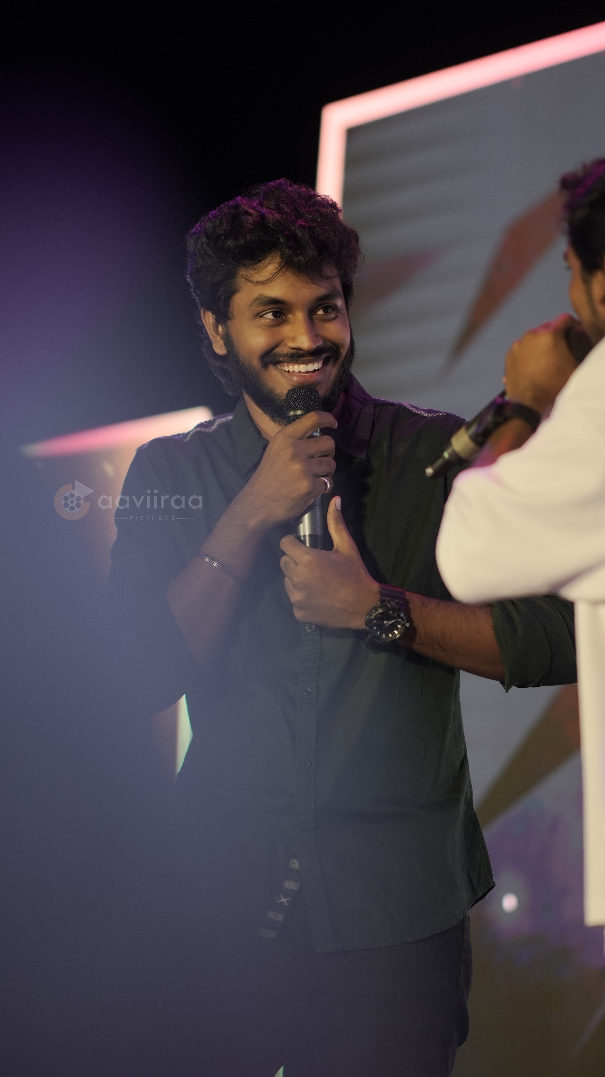 A man with dark, curly hair and a beard smiling while holding a microphone. He is talking to and interacting with another person, who is partially visible. The background is dark with some colorful lighting and a screen.